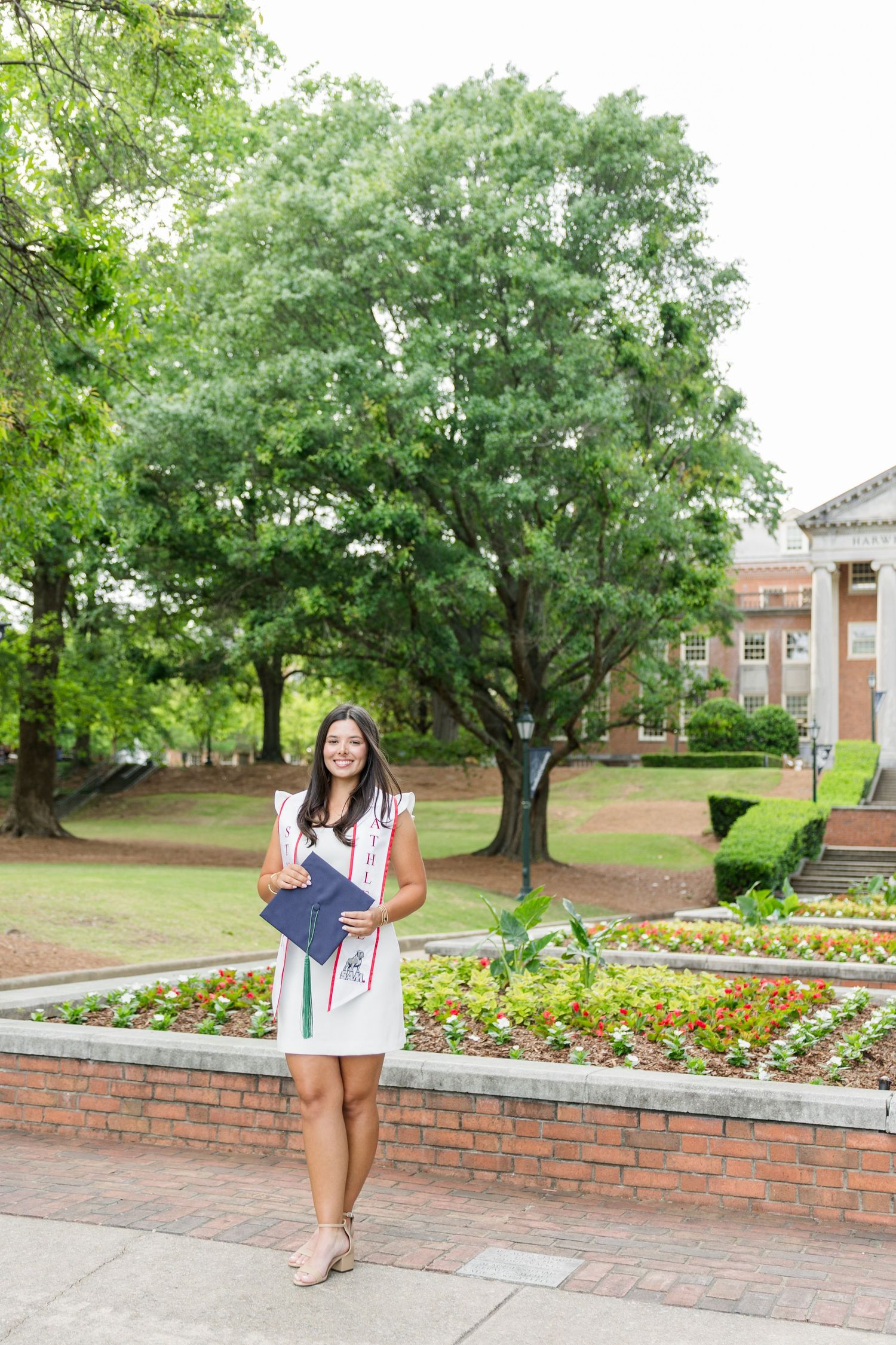 Smiling graduate in white dress holds diploma on a scenic college campus with lush greenery and brick buildings.
