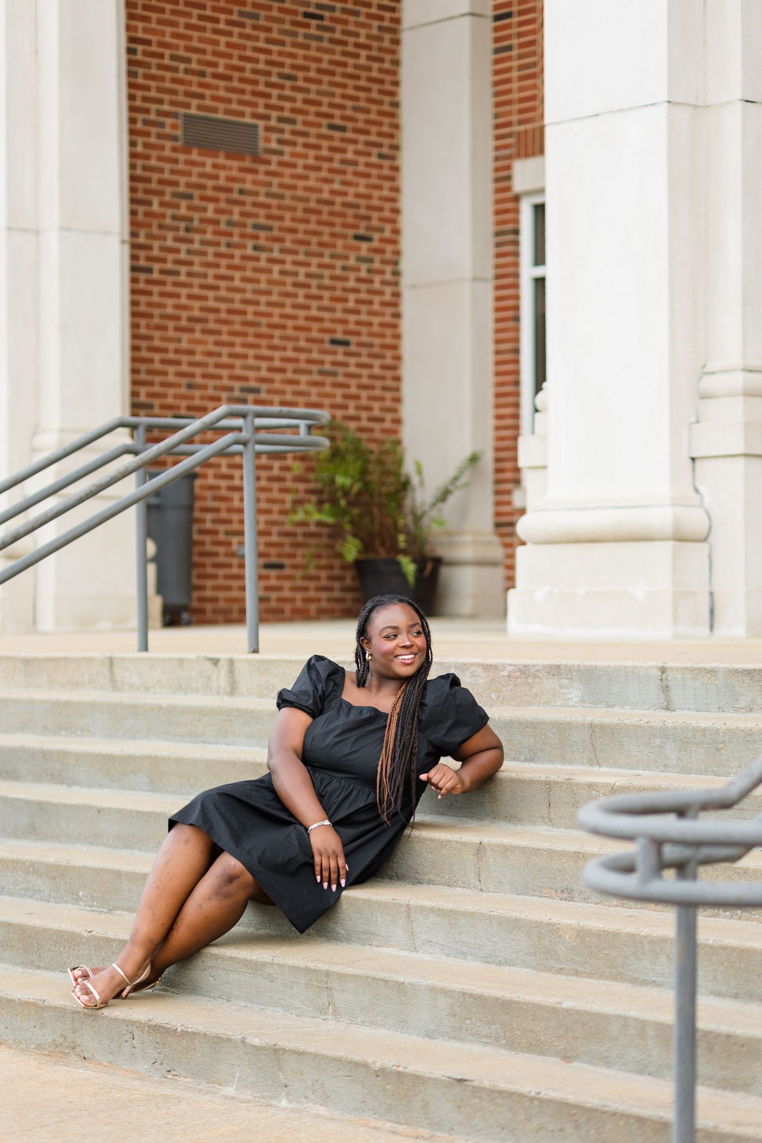 A smiling young Black woman in a black dress sits on stone steps outside a brick building.