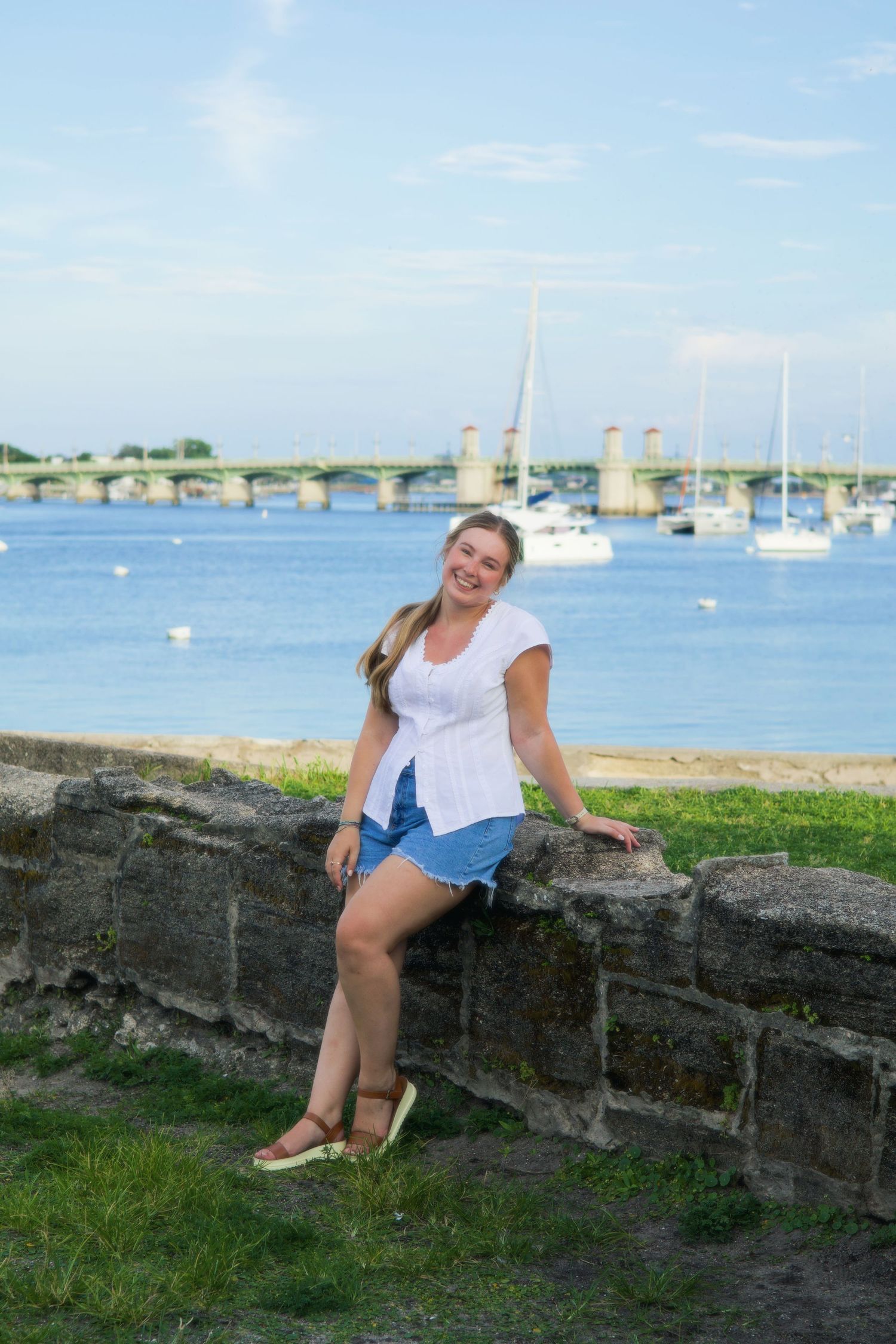 Smiling young woman sitting on stone wall with sailboats and bridge in background.