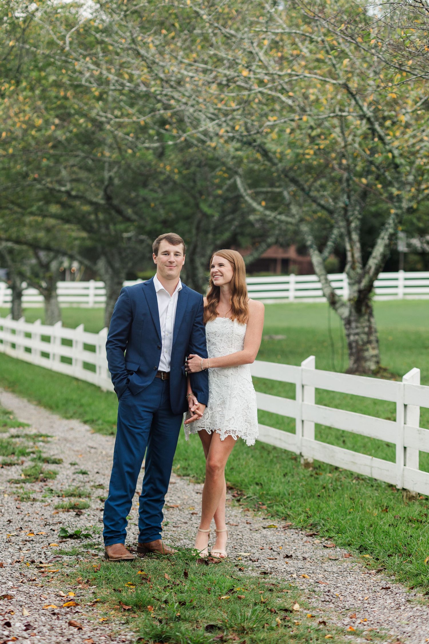 Engaged couple posing together on a scenic gravel path beside a white fence surrounded by trees.