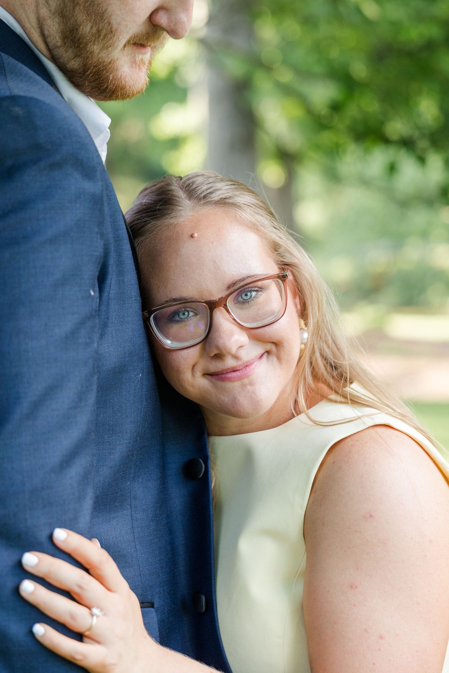 A smiling woman with glasses in a yellow dress leans against a man in a blue suit outdoors.