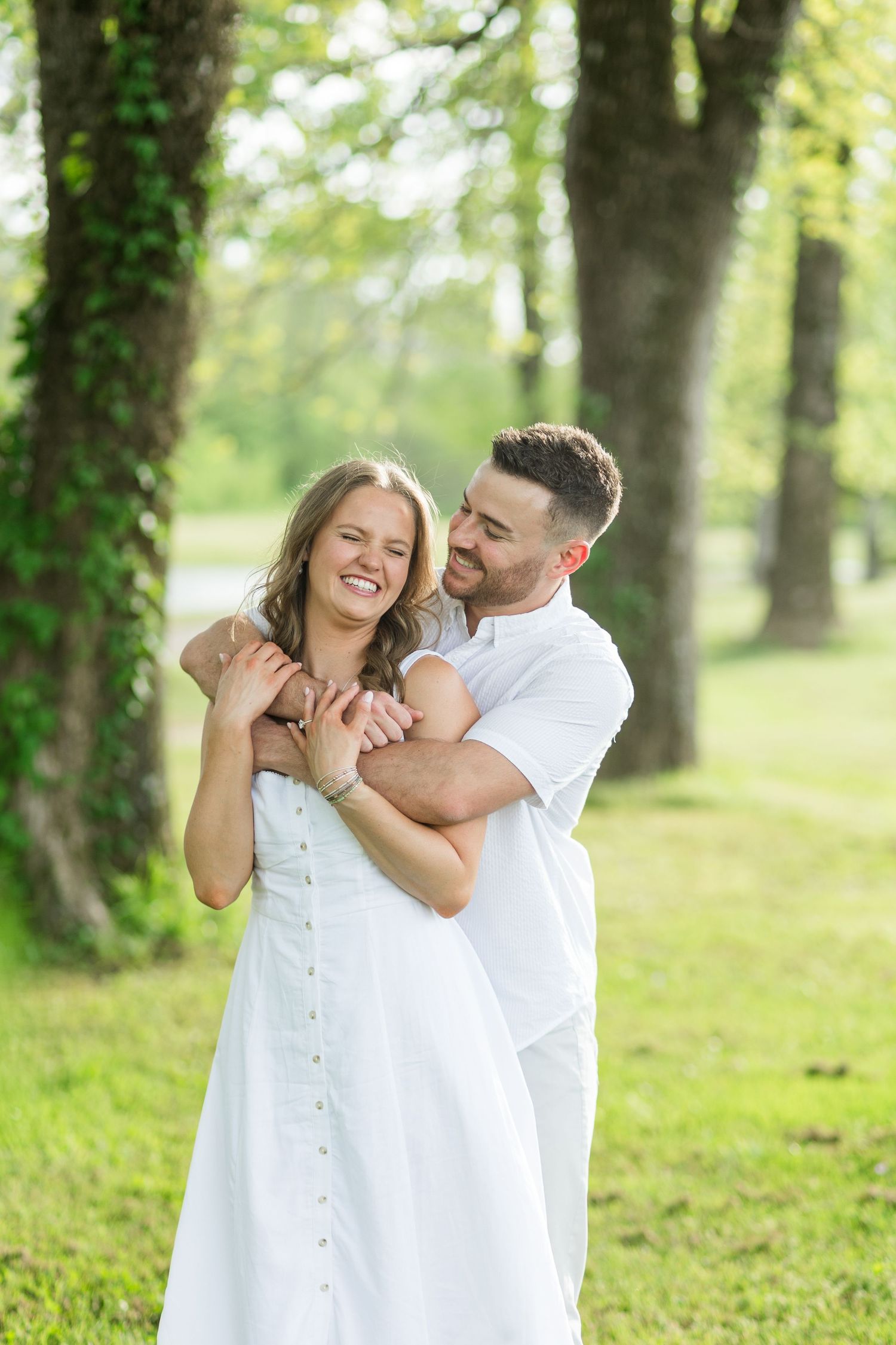 Happy couple embracing outdoors in a lush green park, woman in white dress smiling as man hugs her from behind.
