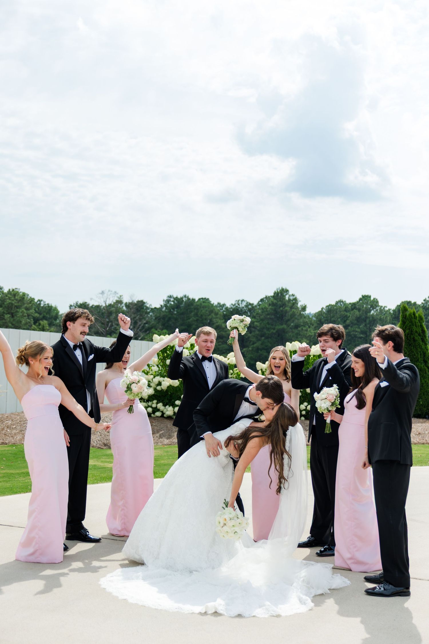 Bride and groom share a dip kiss surrounded by cheering wedding party in pink gowns and black tuxedos outdoors.