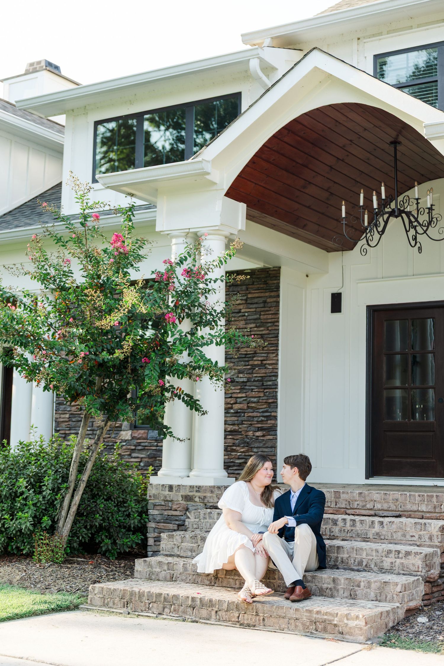Couple sitting on brick steps of elegant white home with stone accents and chandelier porch.