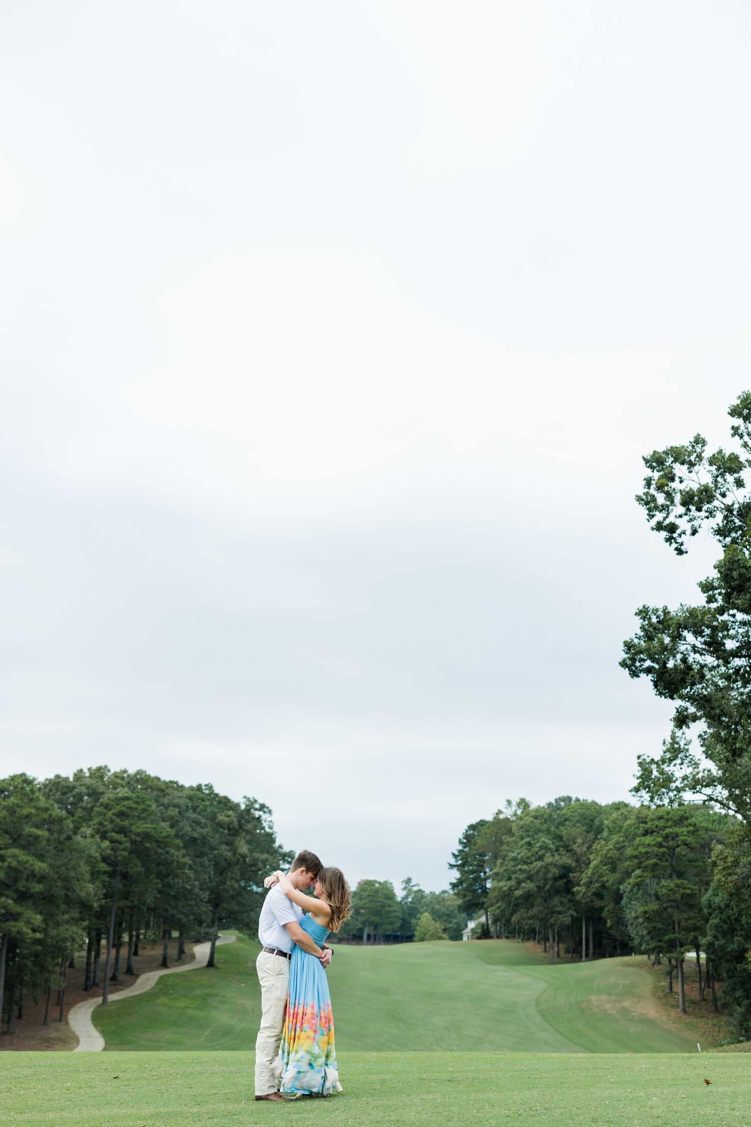 Couple embracing on a golf course fairway in a colorful sundress and khakis, surrounded by tall trees and a winding cart path.
