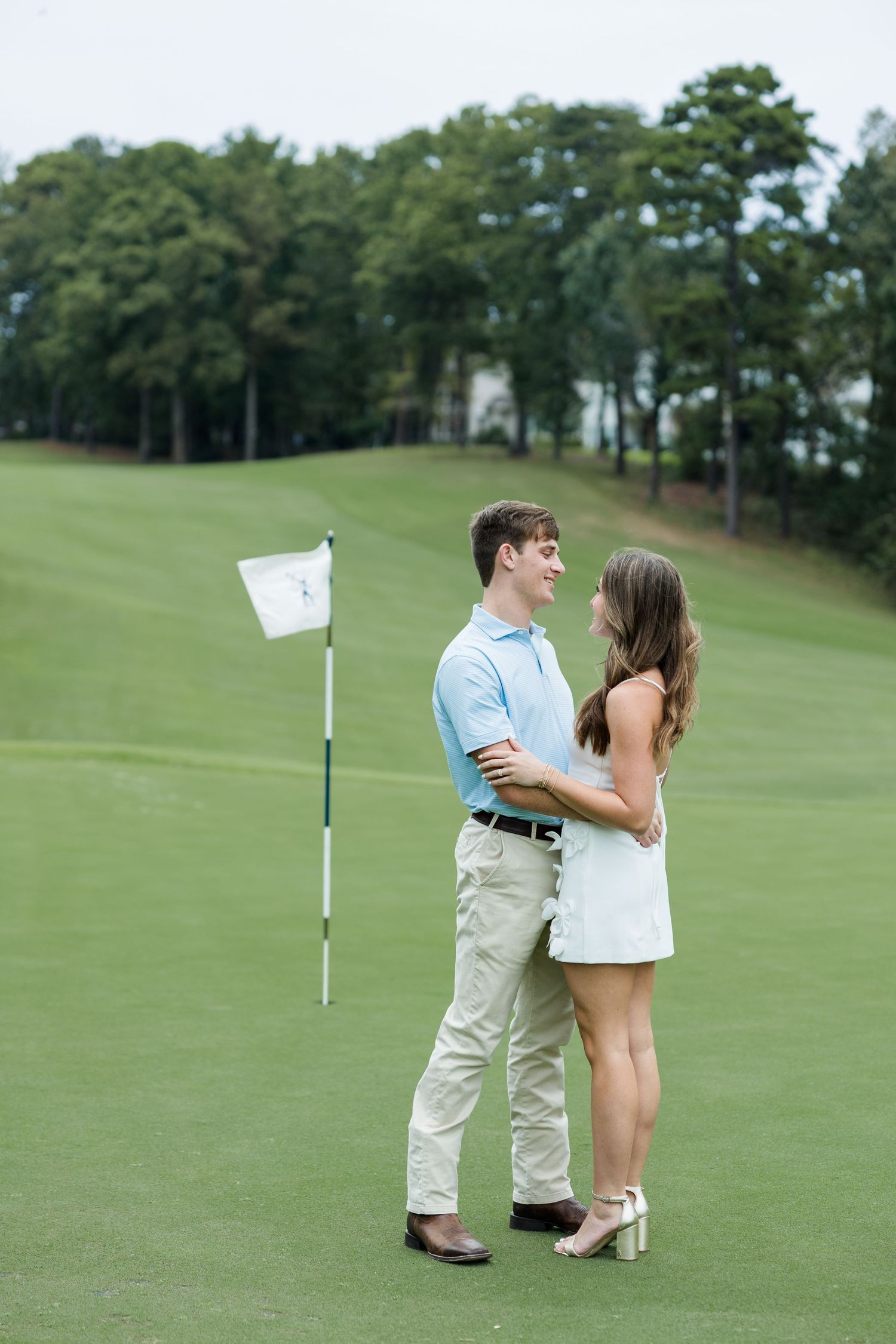 Couple embracing and smiling on a golf course green beside a flag, with tall pine trees behind them in an engagement photo.