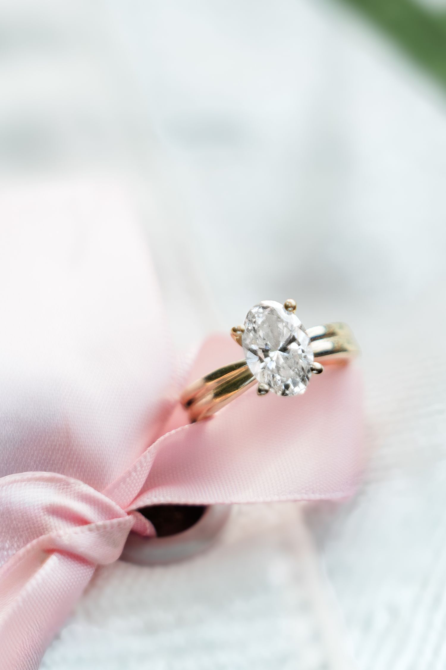 Oval solitaire diamond engagement ring in yellow gold setting displayed on soft pink ribbon and white fabric.