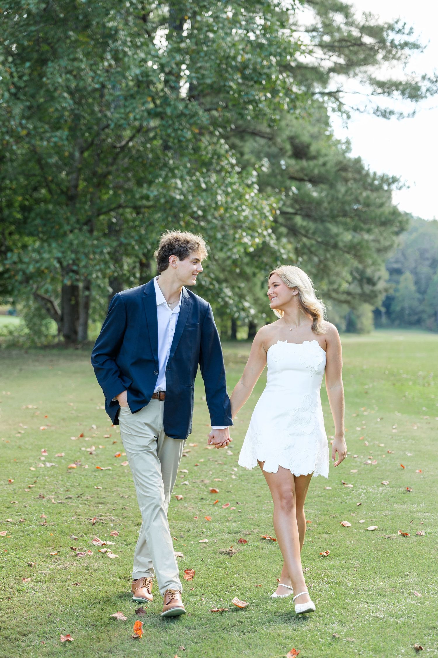 Smiling couple holding hands walking across a lush green golf course, man in navy blazer, woman in white dress.