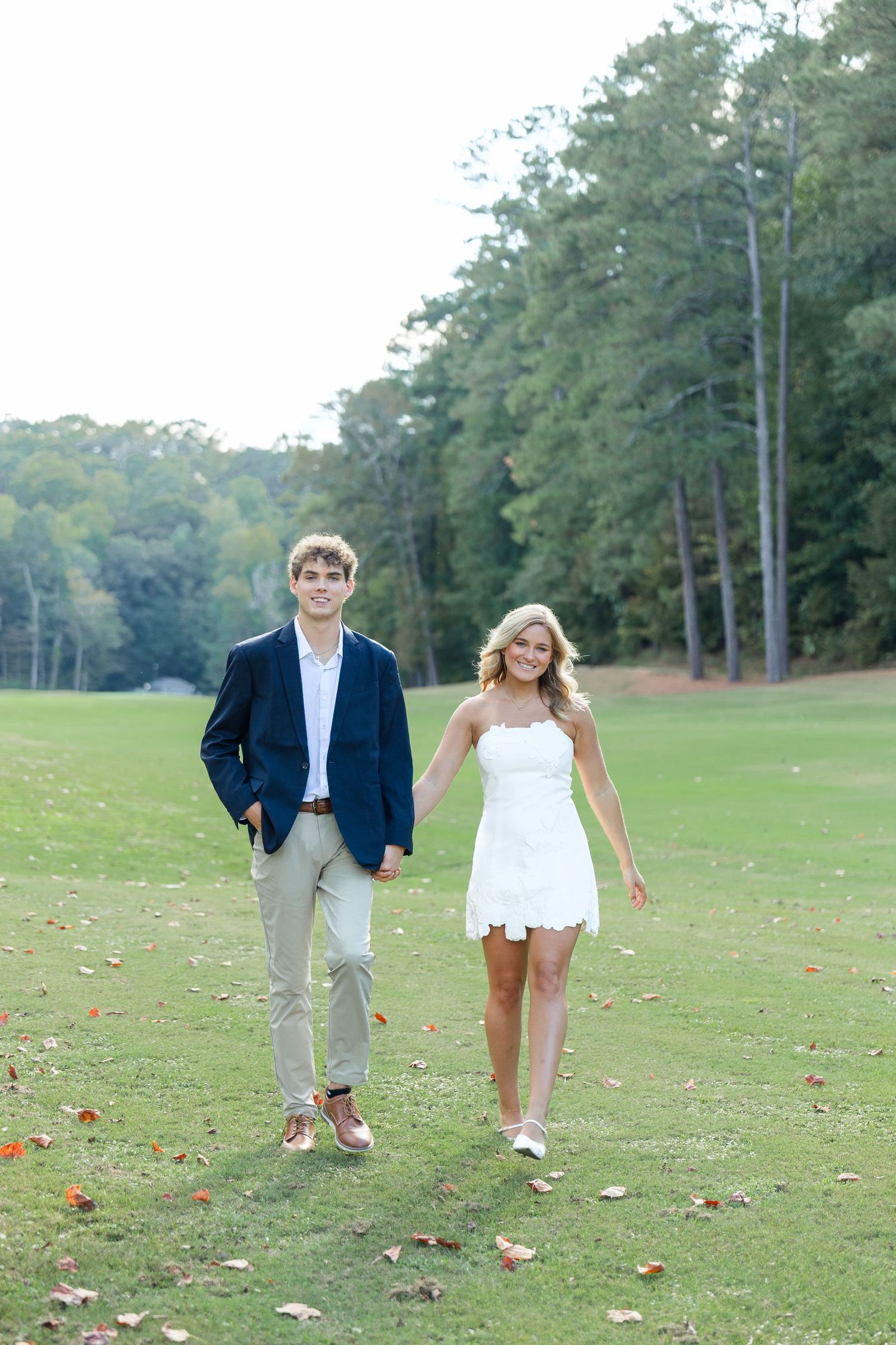 A young couple walks hand-in-hand on a green lawn, man in navy blazer and woman in white dress, with trees behind.