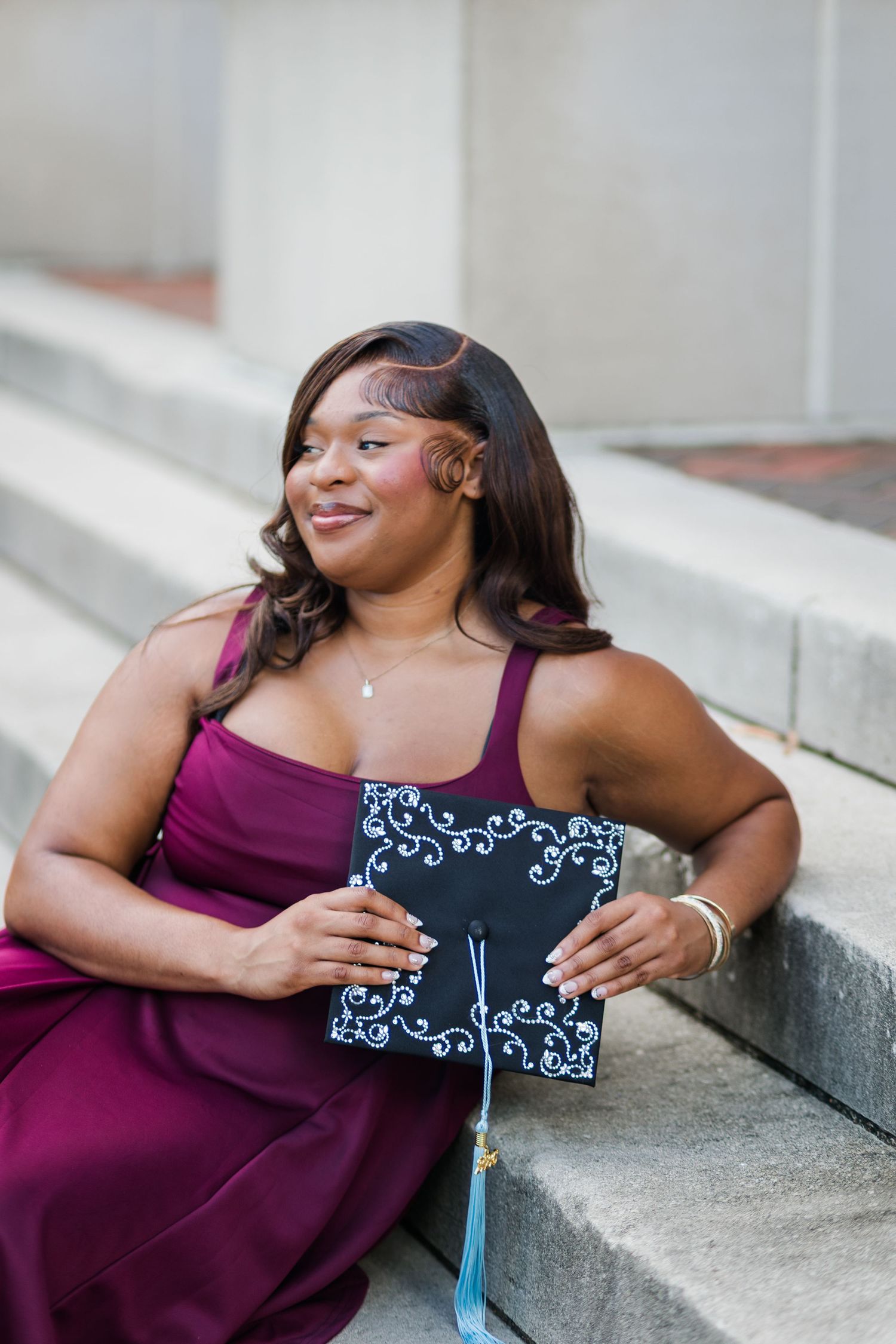 Graduate in burgundy dress sitting on steps holding decorated black mortarboard cap with blue tassel.