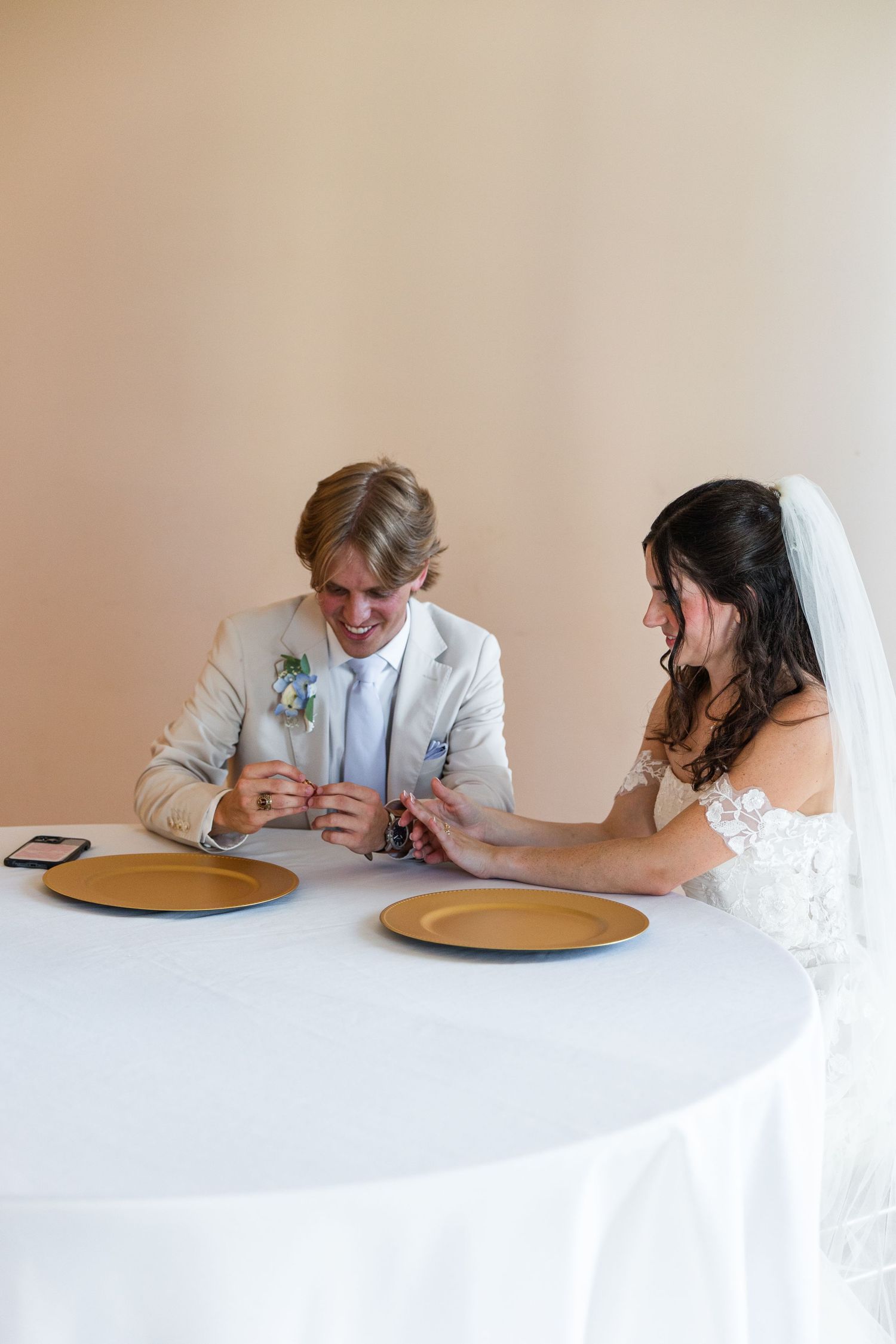 Bride and groom sitting at a white-draped table with gold plates, smiling and exchanging rings at their wedding reception.