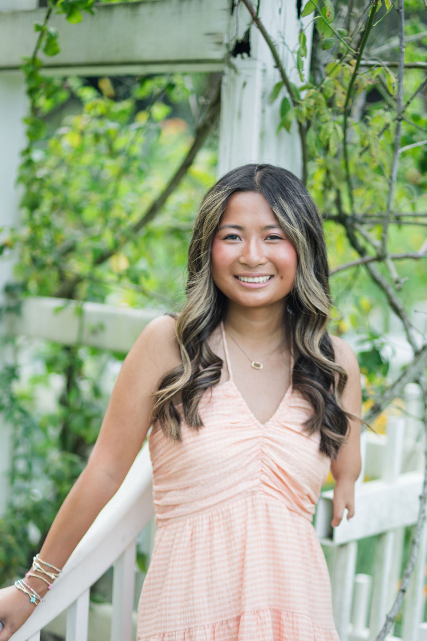 Smiling young woman in peach dress standing by white garden trellis with greenery.