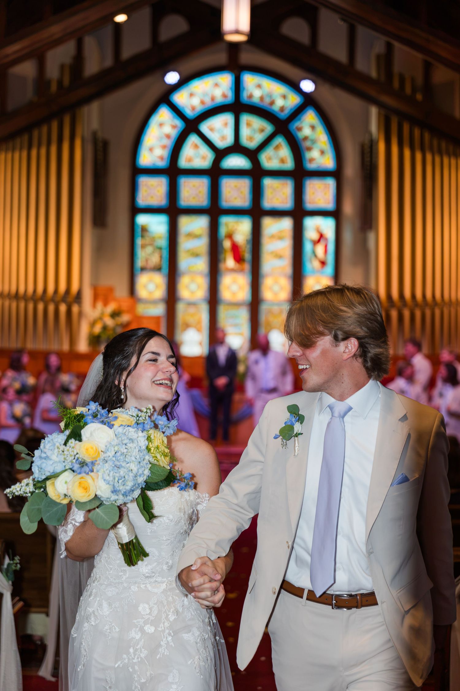 Joyful newlyweds walk down church aisle holding hands, bride carrying blue and white hydrangea bouquet.