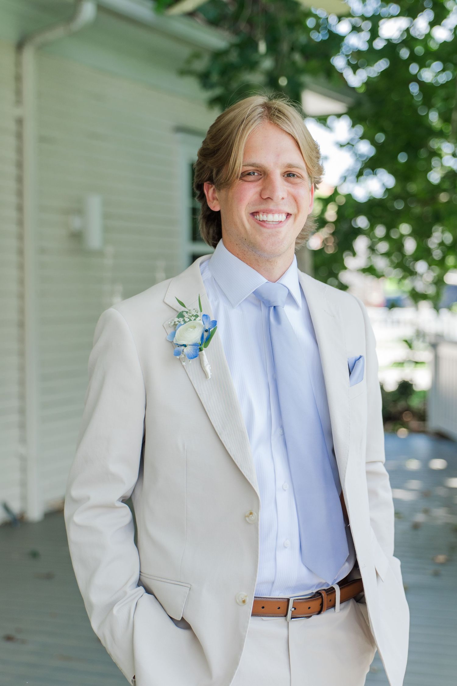 Smiling groom in cream suit with blue tie and floral boutonniere posing outdoors on a sunny wedding day.
