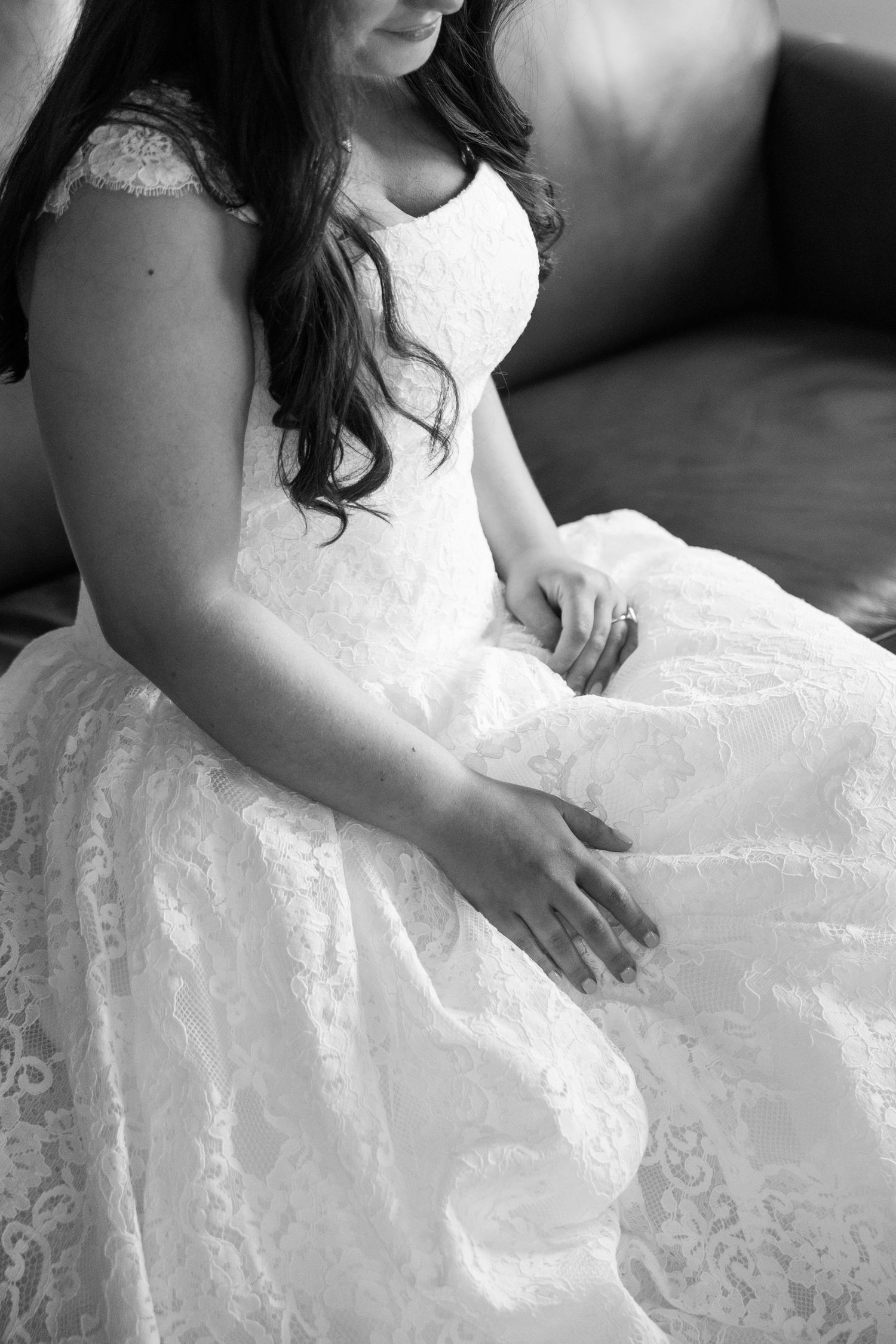 Black and white photo of a bride in a lace wedding gown seated on a sofa, hands resting gently on her dress.