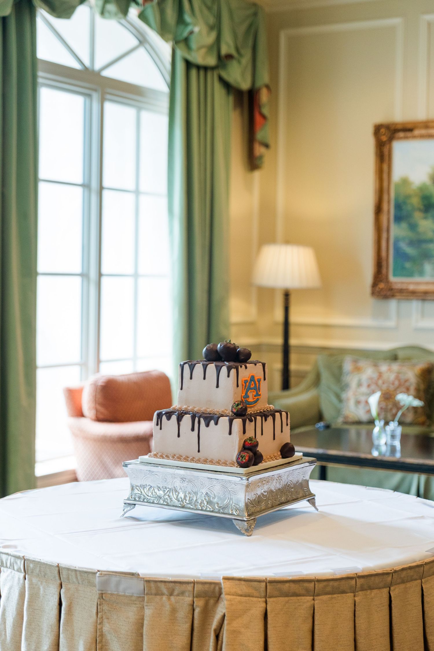 Two-tier white wedding cake with chocolate drip and fresh berries on silver stand in elegant room.