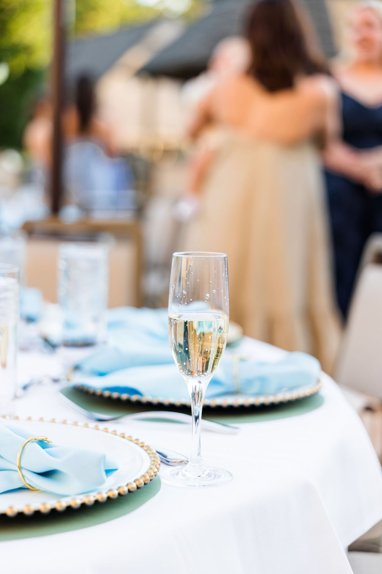 Elegant outdoor wedding reception table set with champagne flute, blue napkins, and gold-rimmed charger plates.