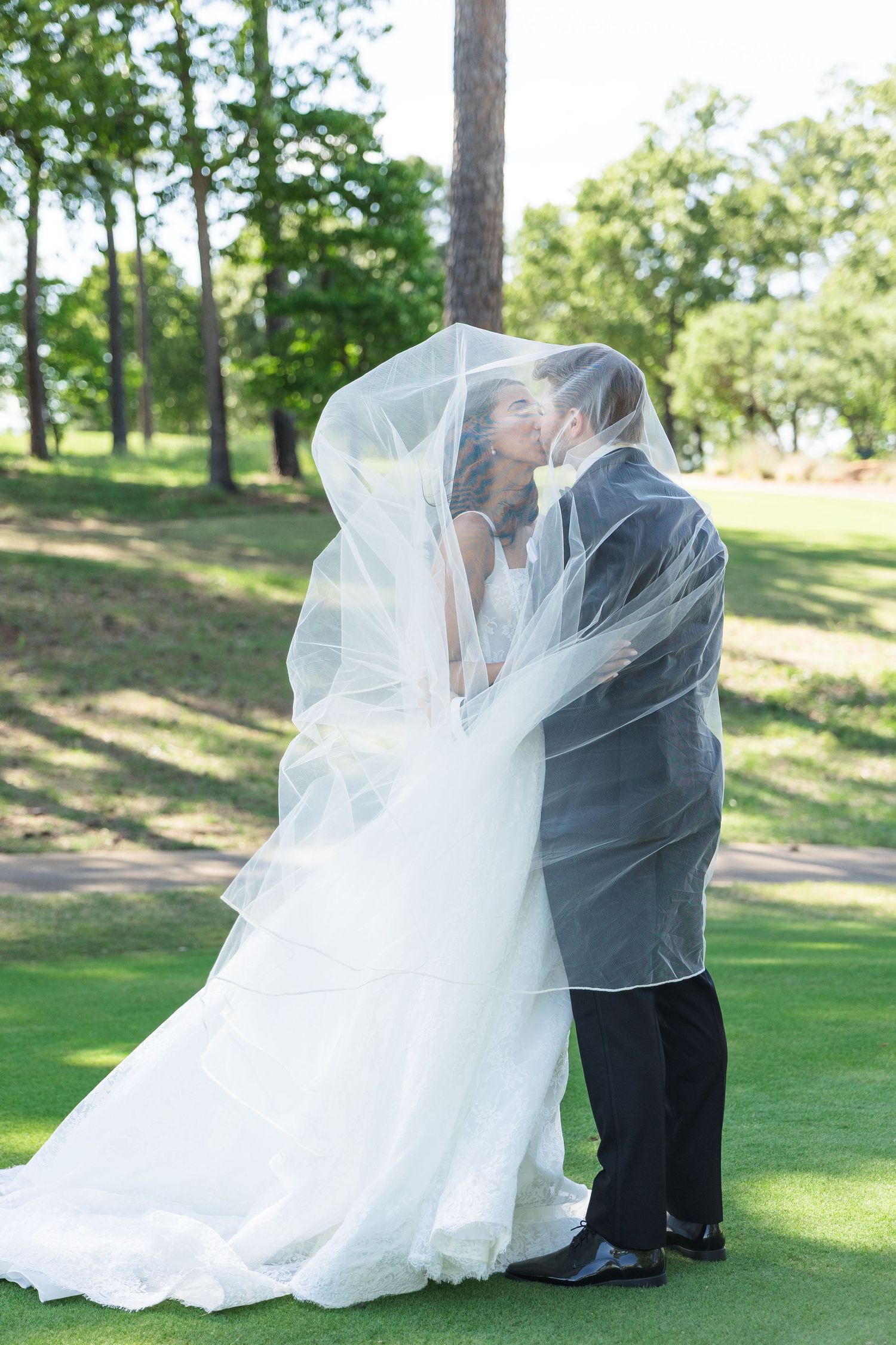 Bride and groom sharing a kiss under a flowing white veil on a lush green lawn surrounded by trees.