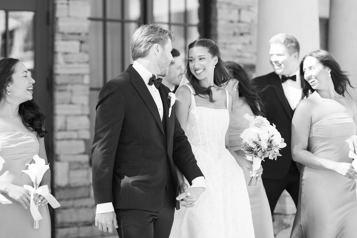 Black and white photo of a smiling bride and groom holding hands, surrounded by wedding party members in formal attire.