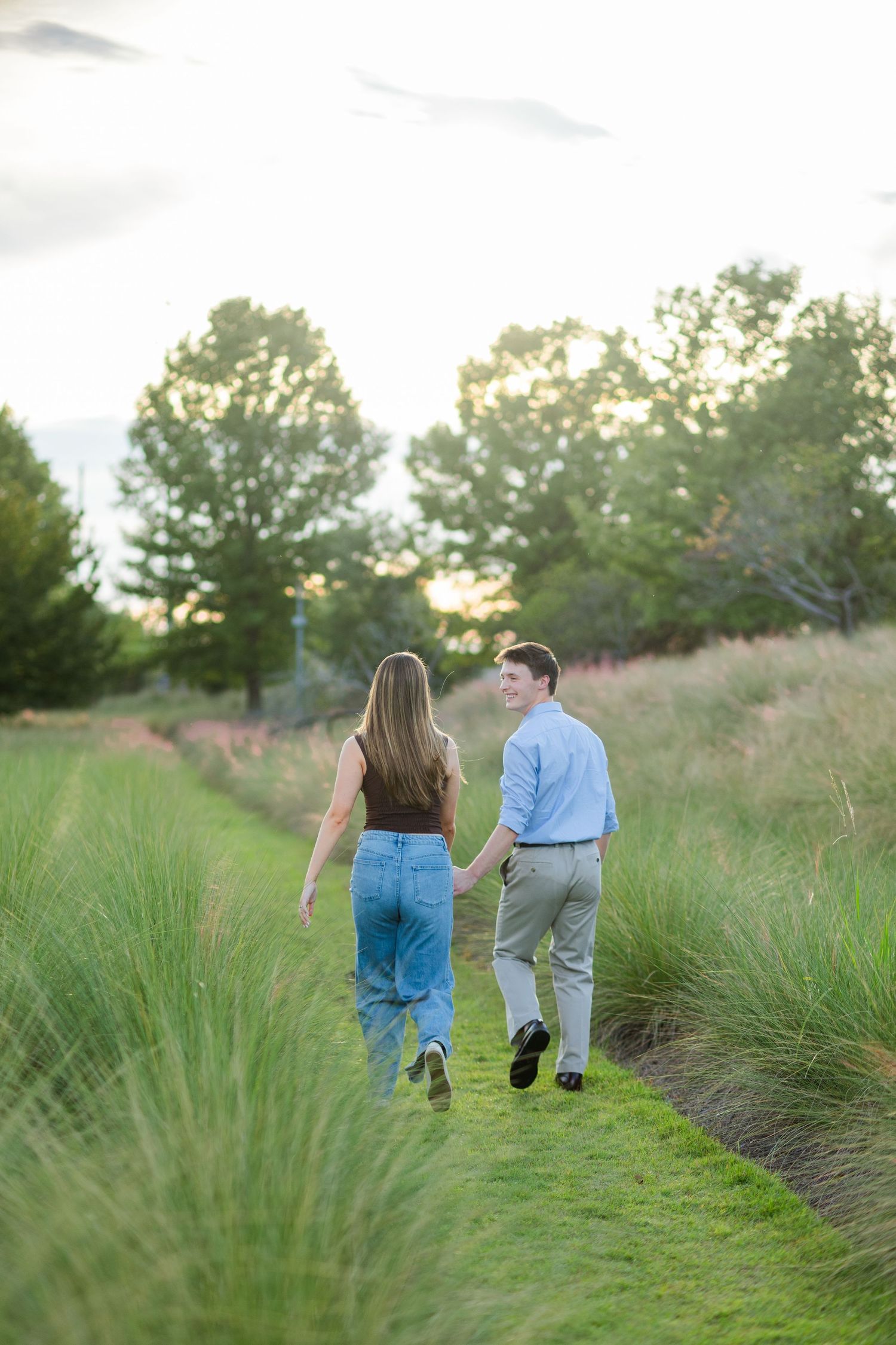 A couple holds hands while walking along a lush green path surrounded by tall ornamental grasses.