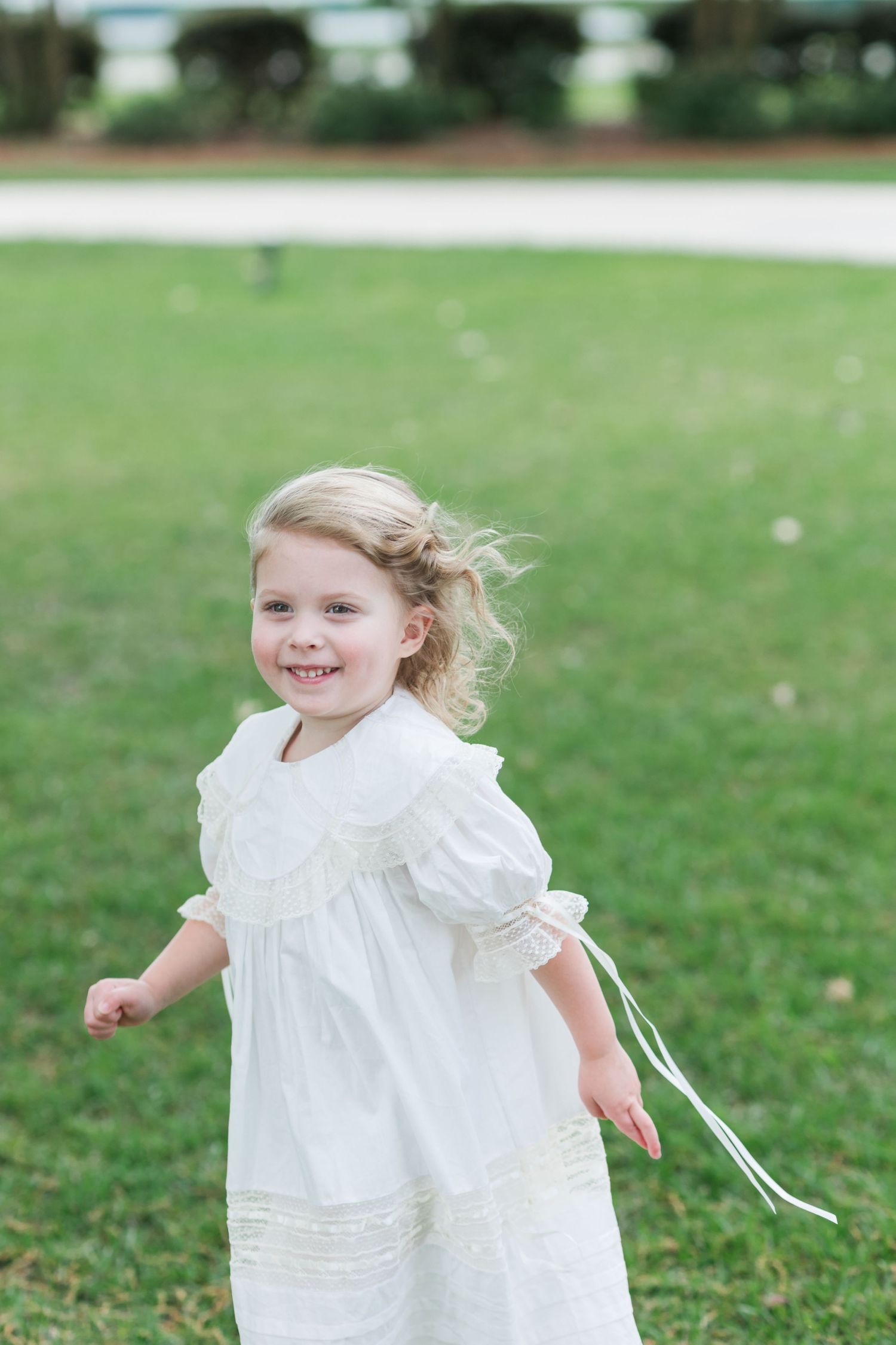 Smiling blonde flower girl in white lace dress runs across green lawn at outdoor wedding.