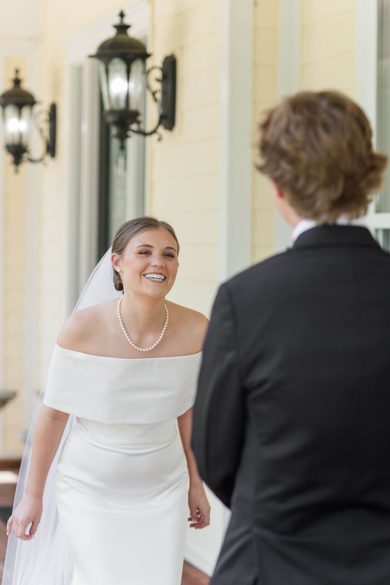 Smiling bride in off-shoulder white dress with pearl necklace shares first look moment with groom.