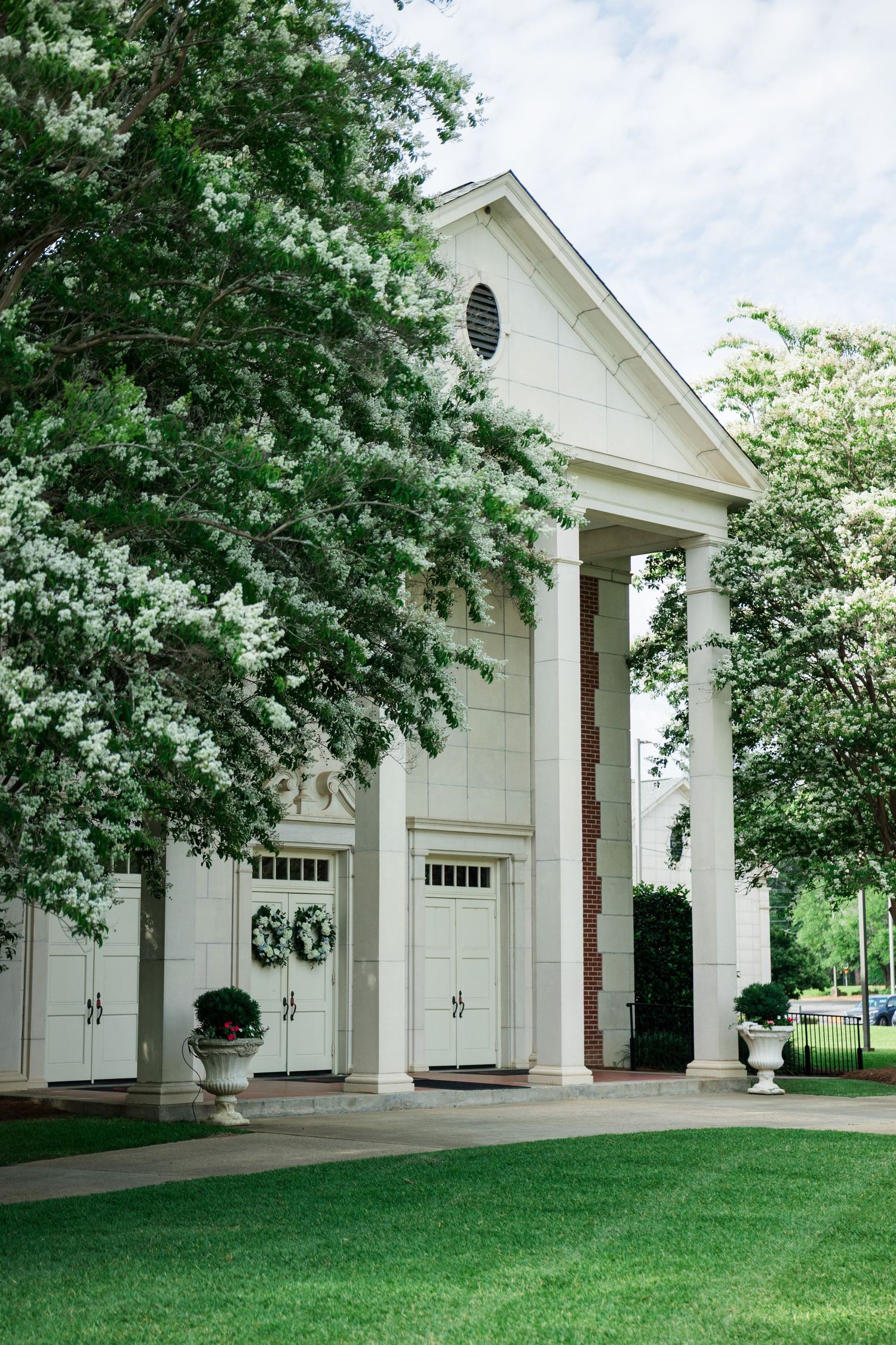 White colonial building with tall pillars, blooming trees, and lush green lawn on a cloudy day.