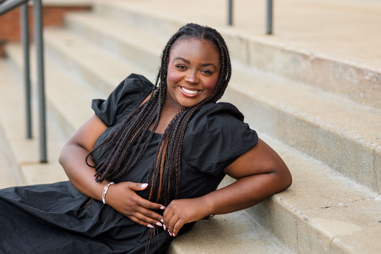 Smiling young woman with braids in black dress poses on outdoor stone steps.