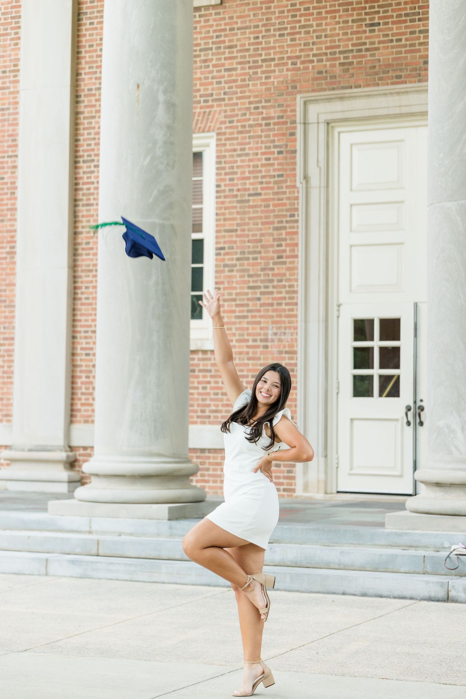 Young woman in white dress tosses graduation cap in front of brick building with white columns.