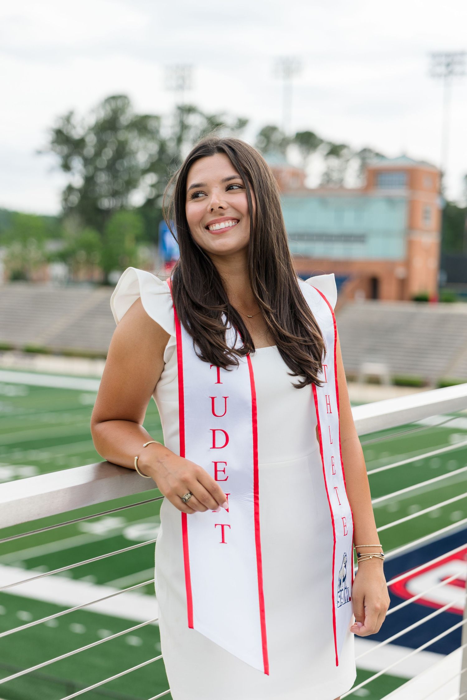 Smiling graduate in white dress and sash poses at stadium railing during outdoor graduation photo session.