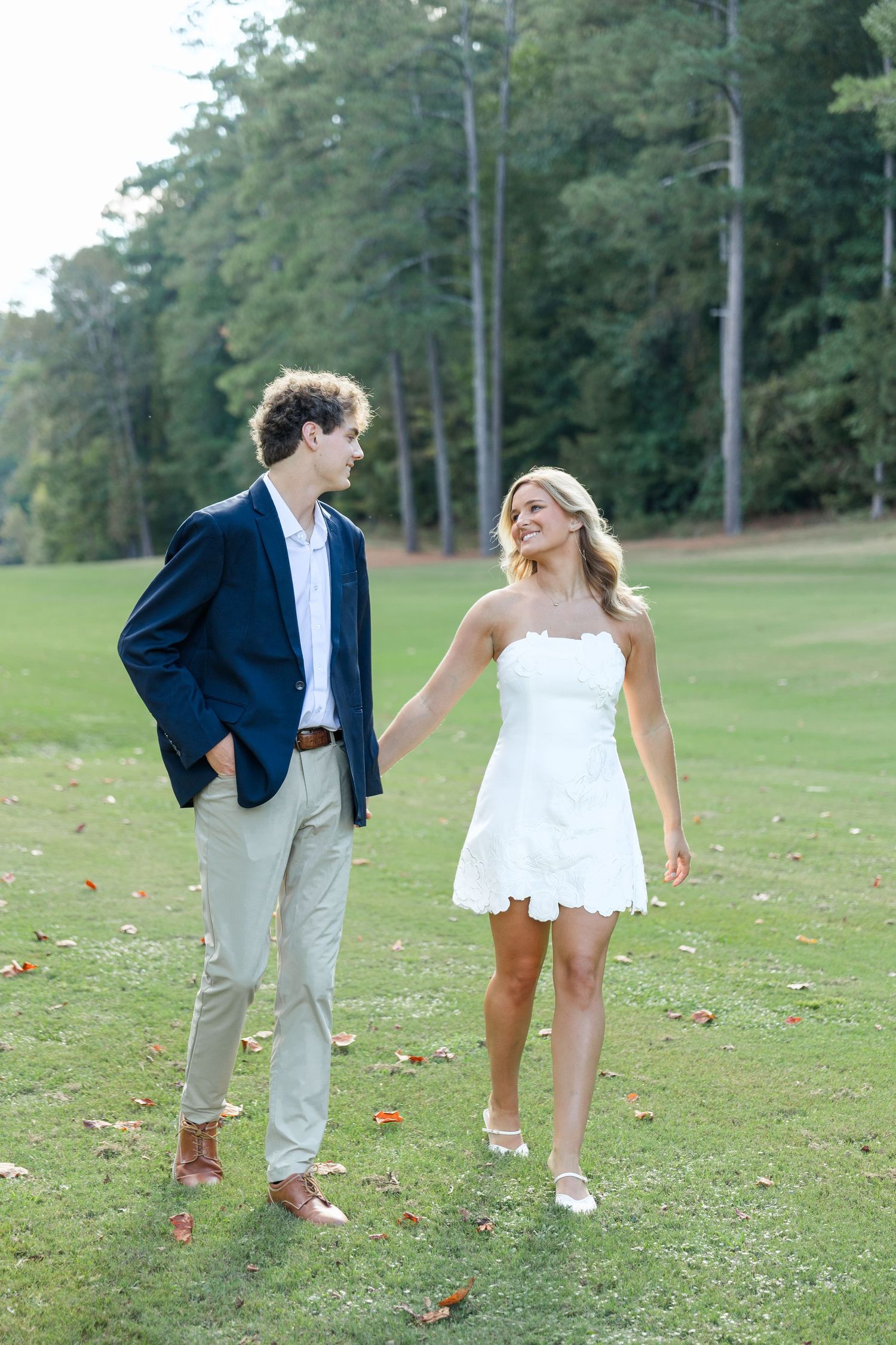 A couple walks hand-in-hand on a green lawn, man in navy blazer and woman in white mini dress, with trees behind.