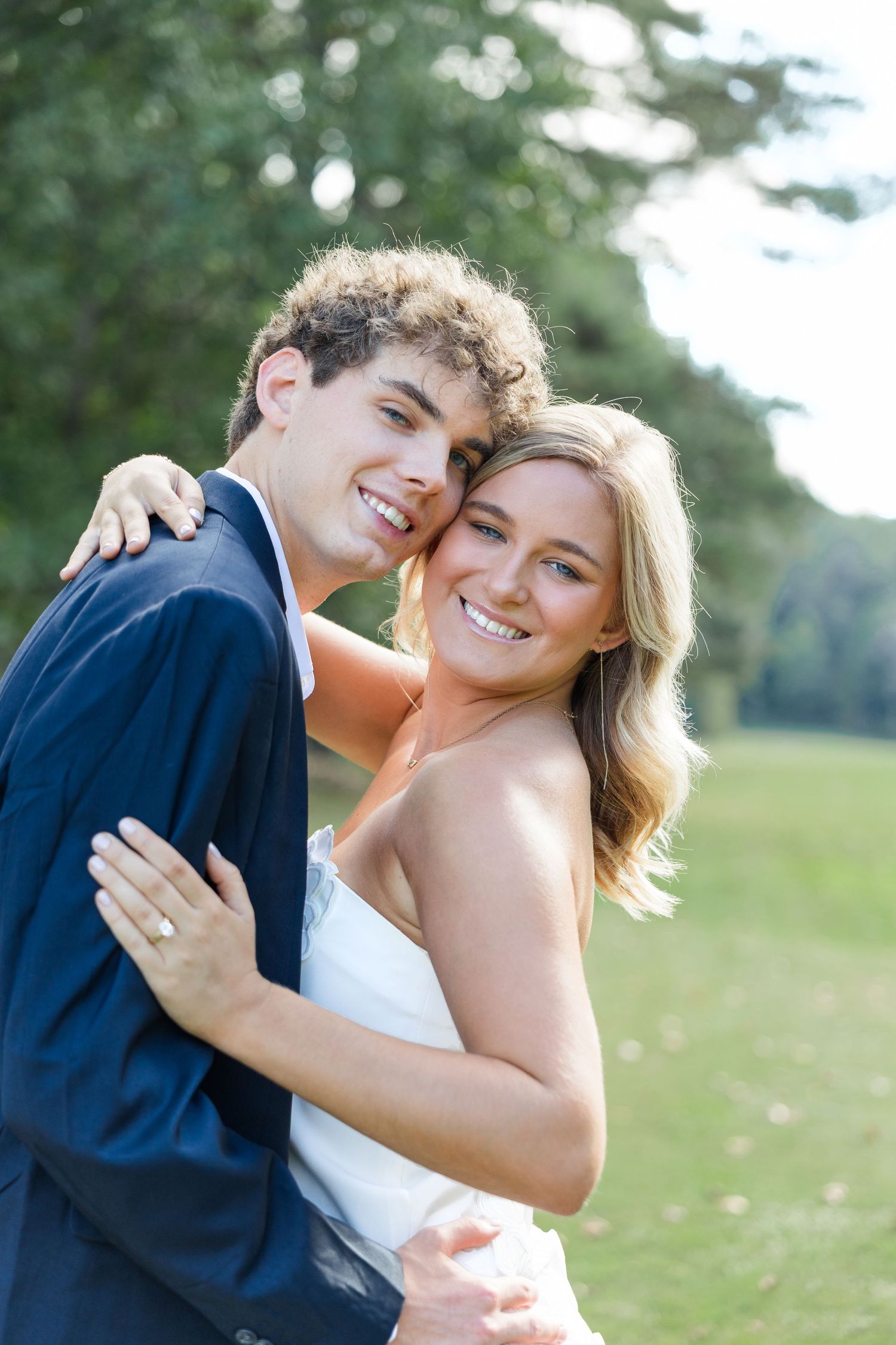 Happy engaged couple embracing outdoors on a golf course, woman showing diamond ring, both smiling at camera.