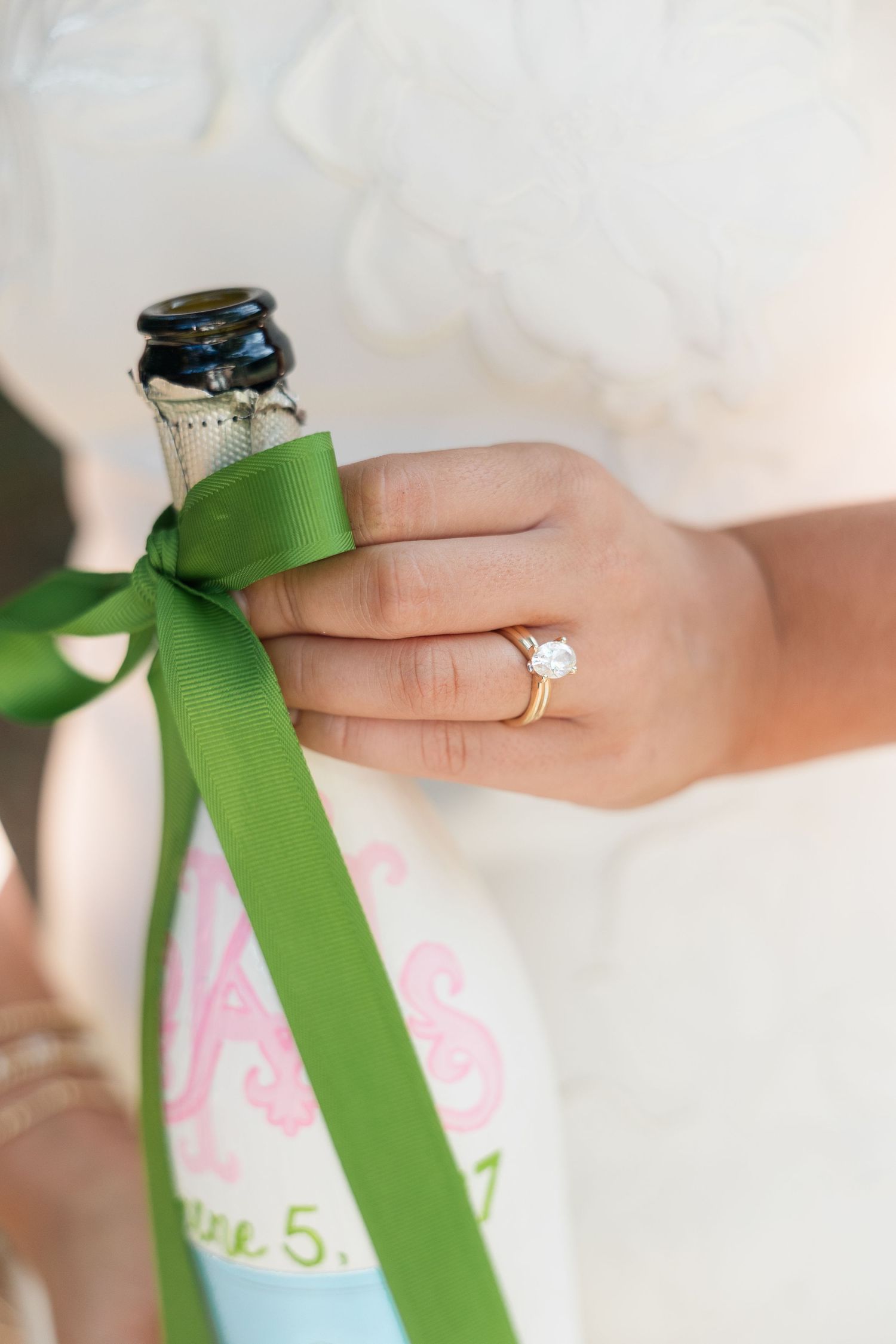 Bride holding champagne bottle with green ribbon, showcasing round solitaire diamond engagement ring on gold band.