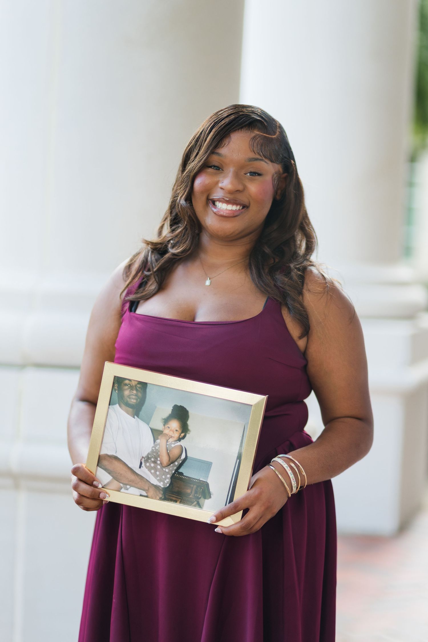 Young woman in burgundy dress smiling while holding a framed photo at a graduation ceremony.