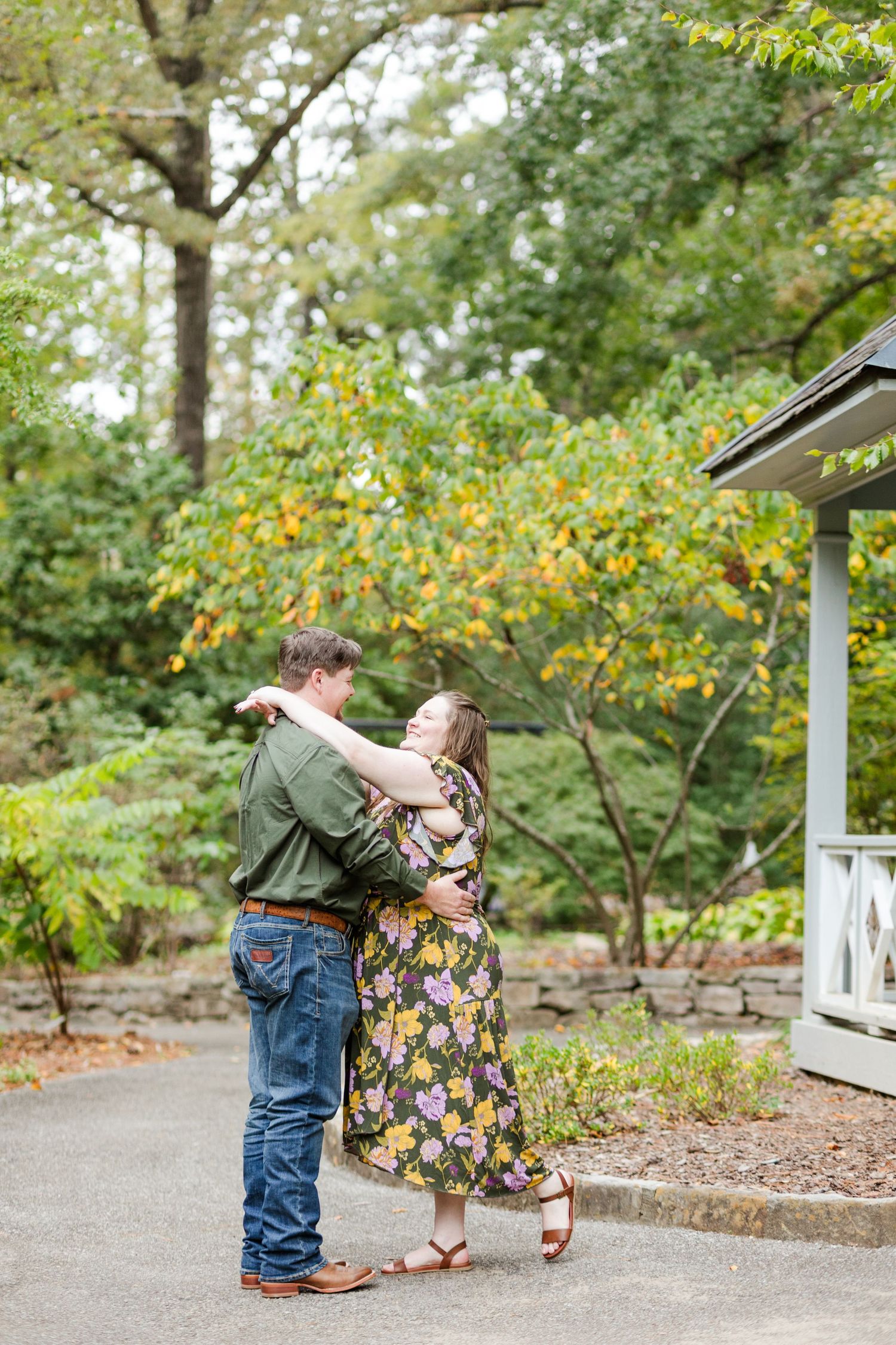 Couple embracing and dancing outdoors in a lush garden, woman in floral dress, man in green jacket and jeans.