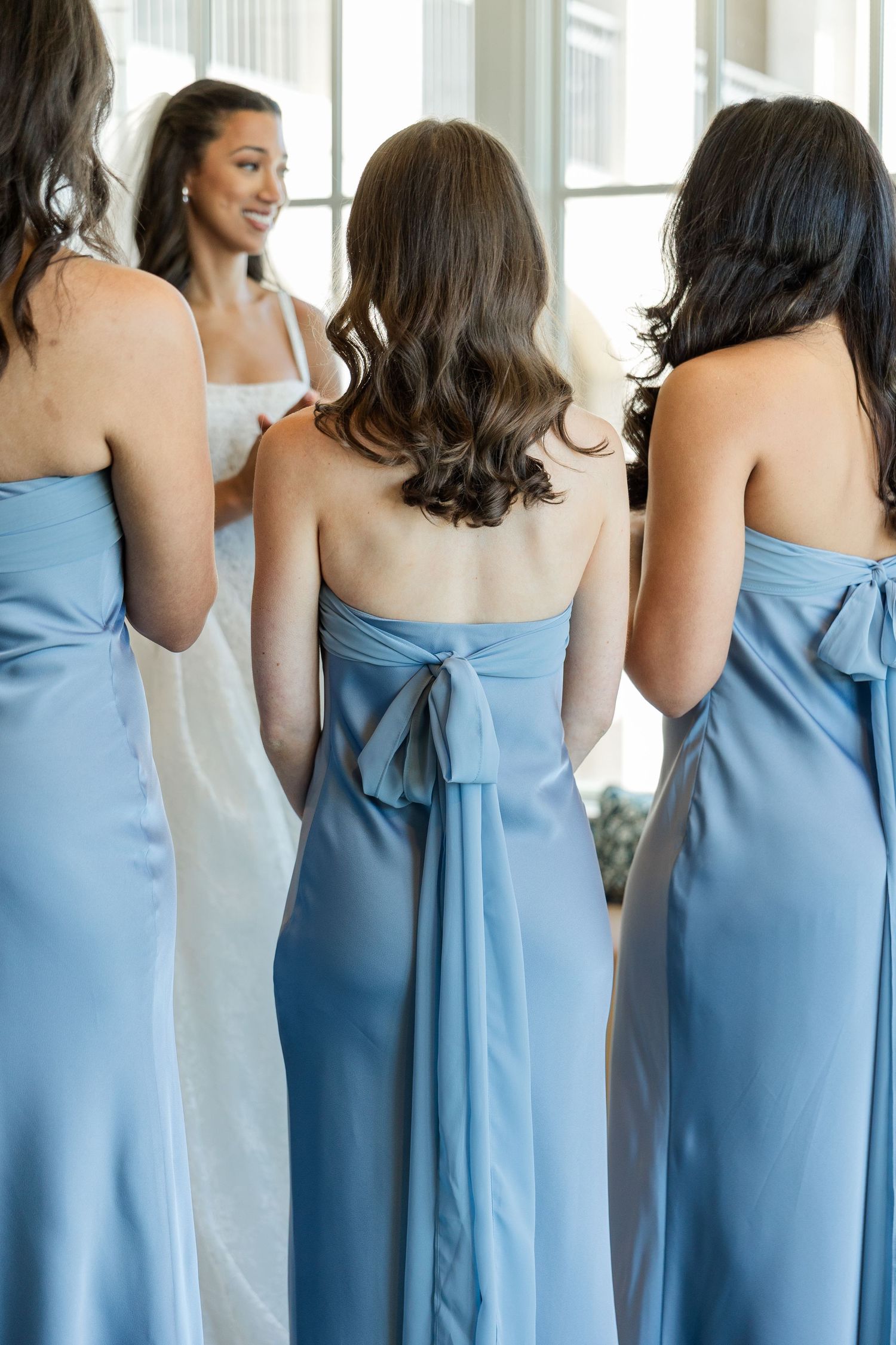 Bridesmaids in strapless dusty blue satin gowns with bow backs stand facing a smiling bride in white.