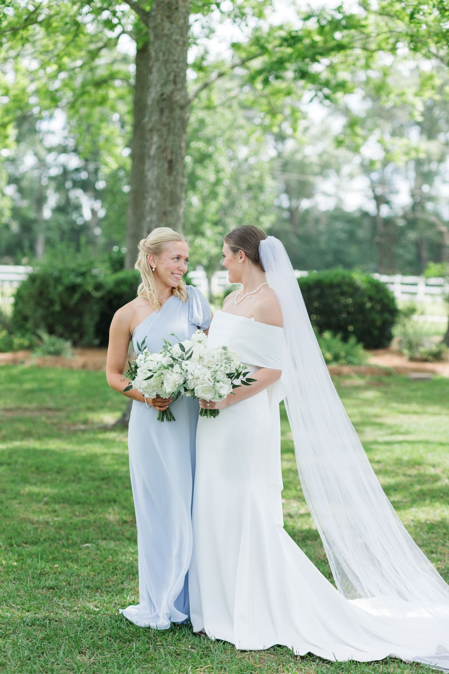 Bride in off-shoulder gown with long veil standing beside bridesmaid in light blue dress outdoors.