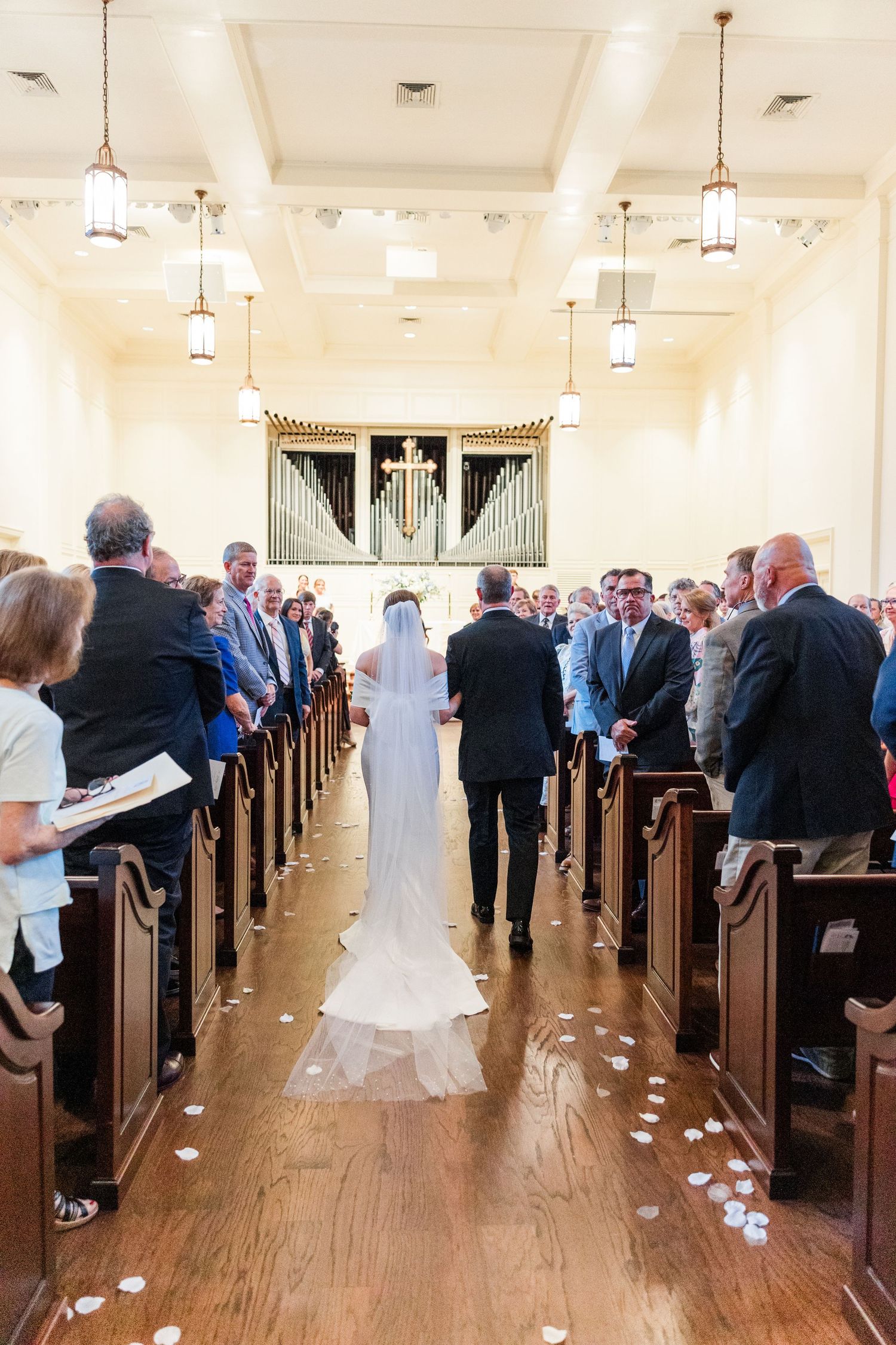 Bride in cathedral veil walks down church aisle with escort past seated wedding guests toward altar.