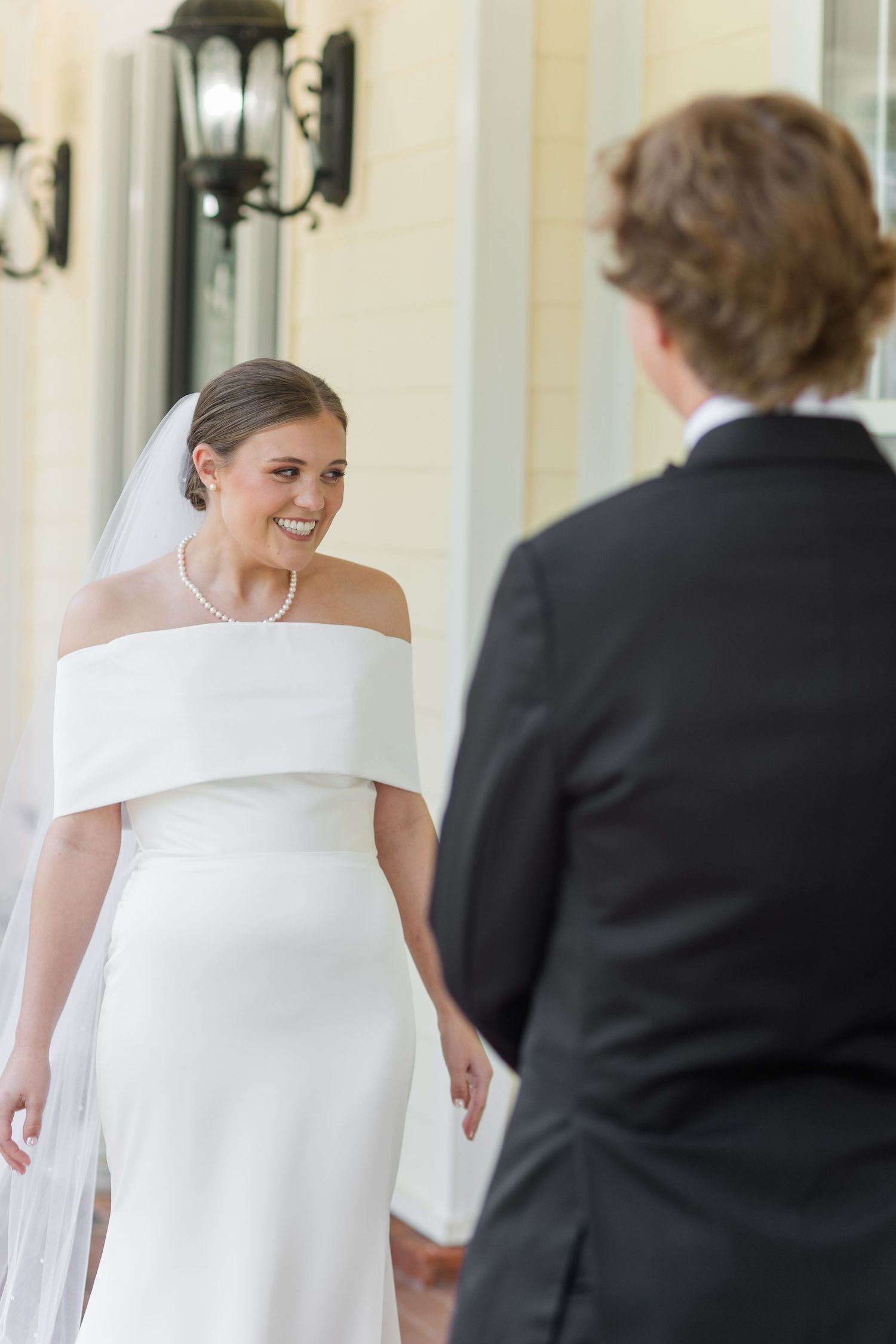 Radiant bride in white off-shoulder gown and veil smiles at groom during outdoor first look.