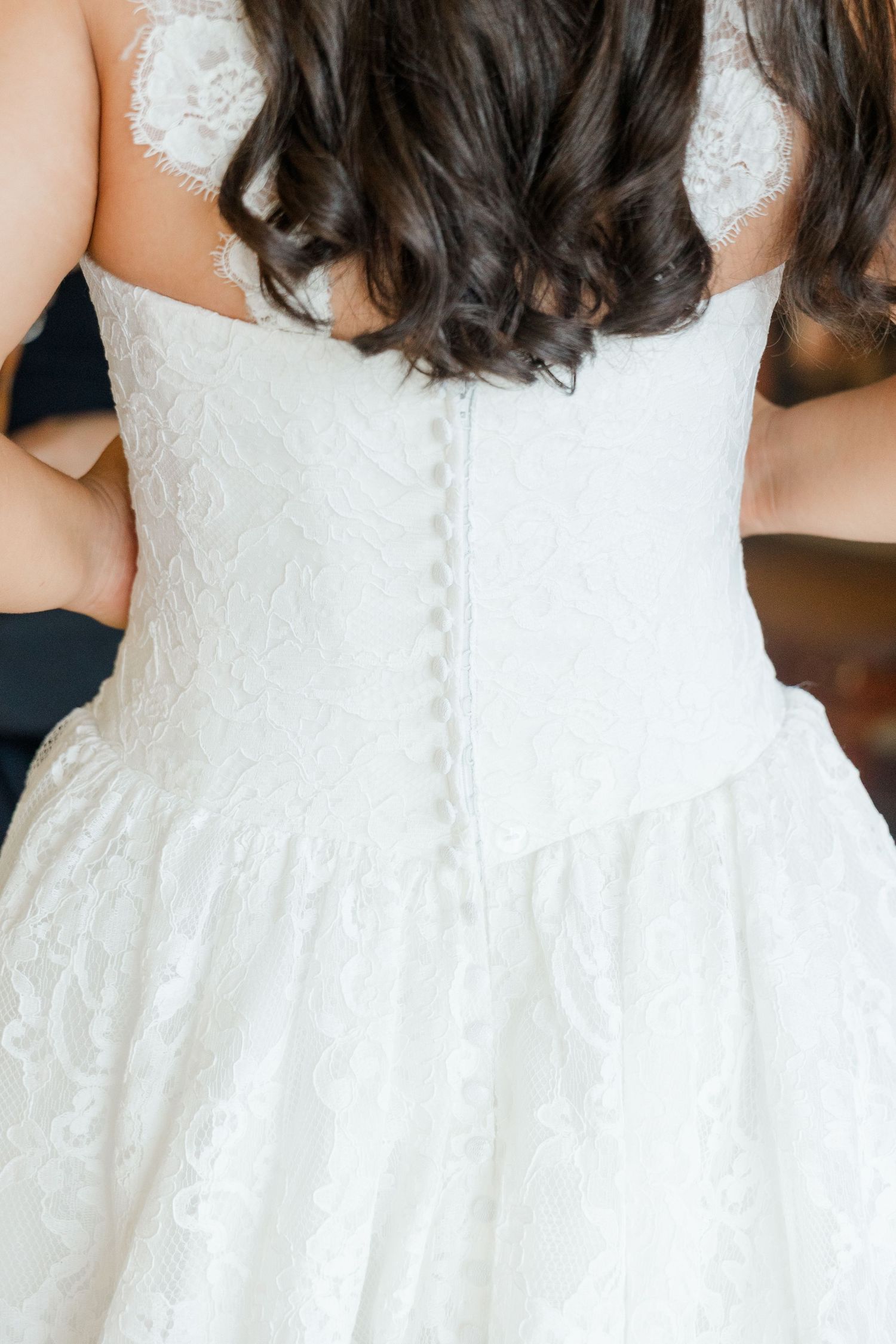 Back view of a bride in a white lace wedding gown with button closure and flowing skirt, dark curly hair.