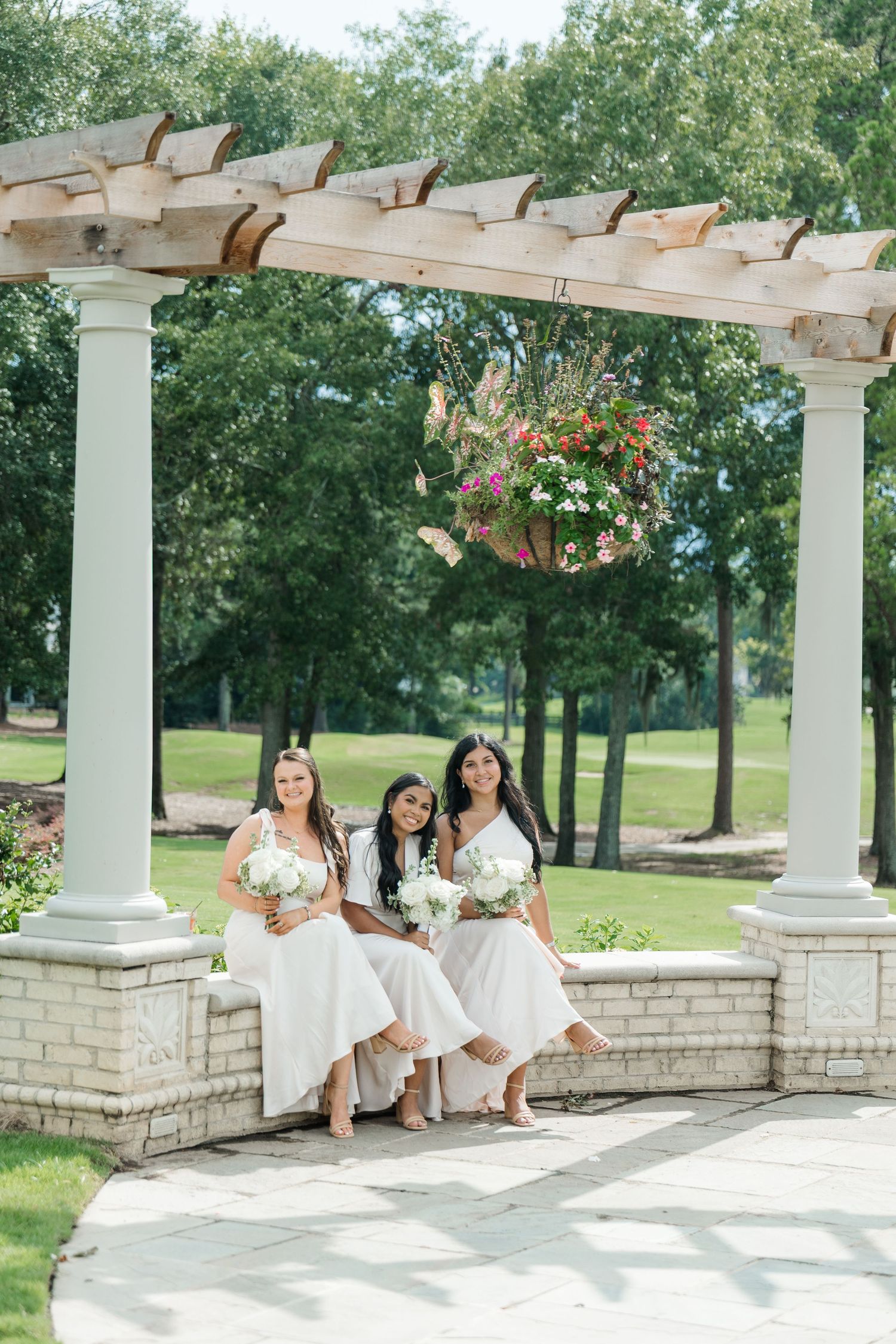 Three bridesmaids in white dresses sit under a pergola with hanging flowers, holding bouquets at an outdoor wedding venue.