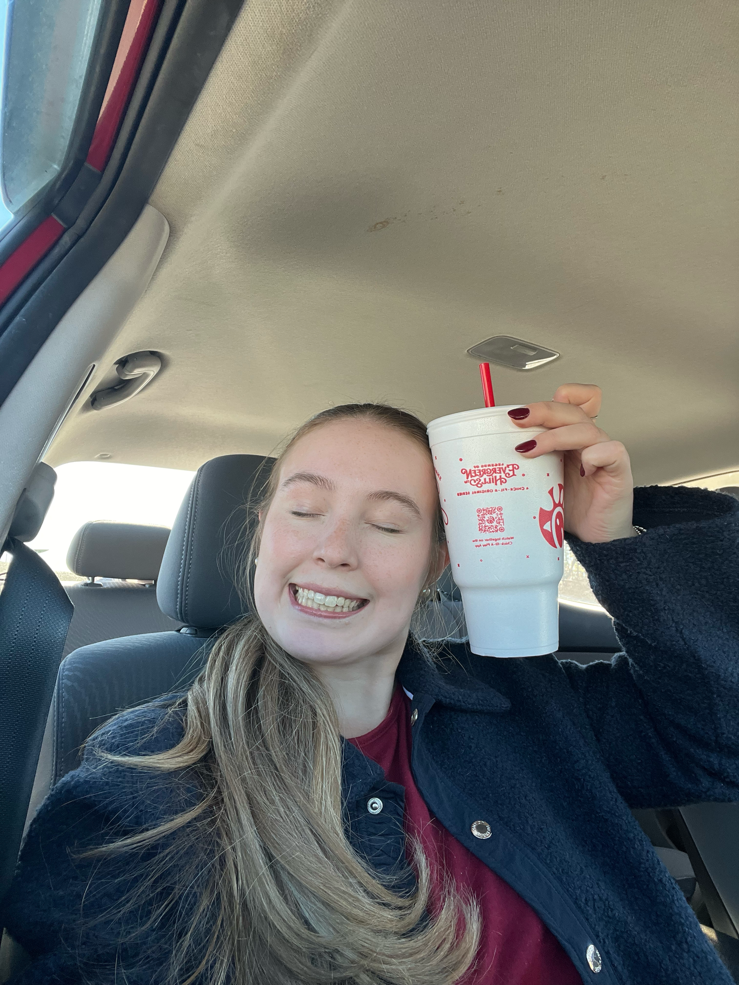 Smiling teenage girl holding a Chick-fil-A cup while sitting in a car, eyes closed happily.