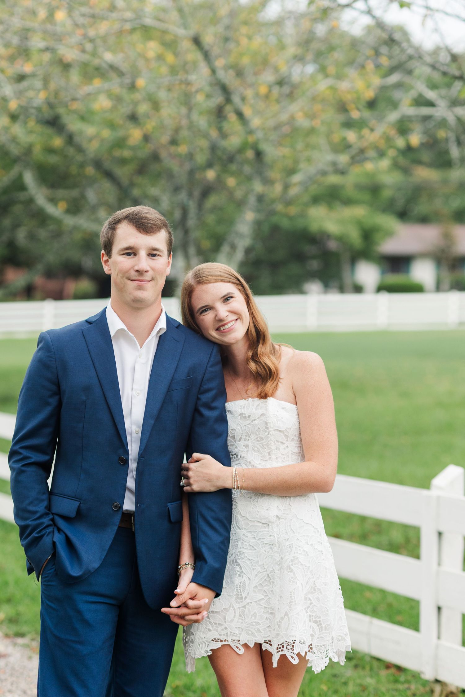 Couple posing outdoors near white fence, man in navy suit, woman in white lace dress, smiling at camera.