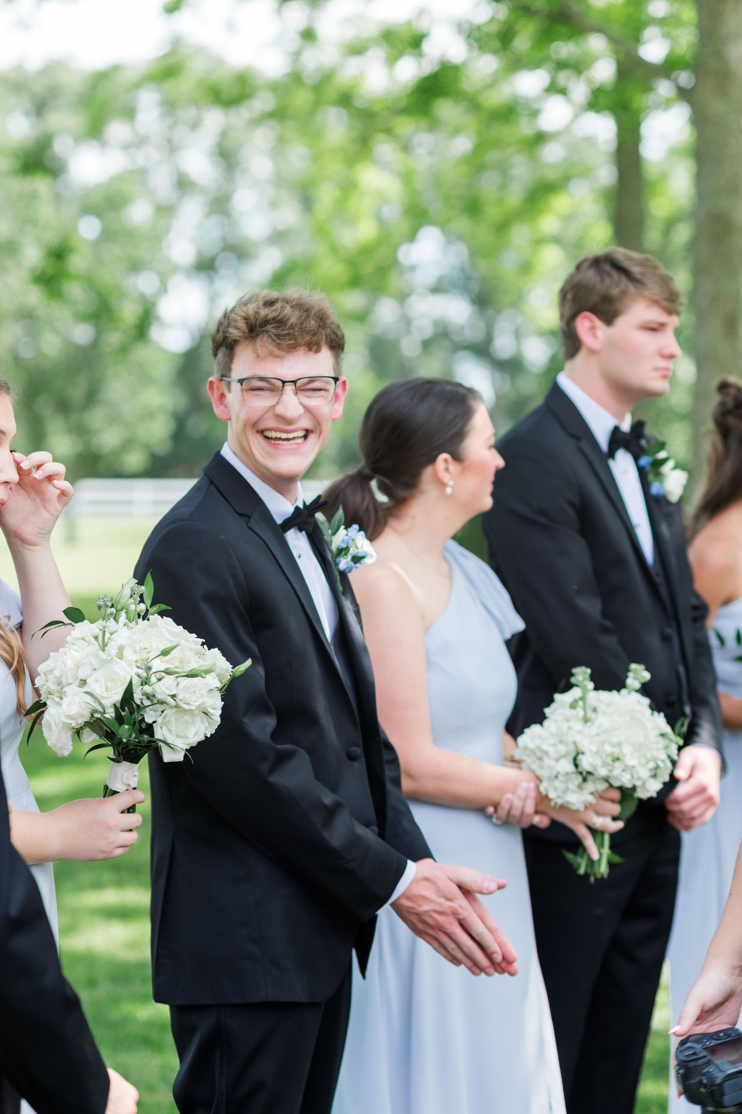 Smiling groomsman in black tuxedo holding white floral bouquet with wedding party outdoors.