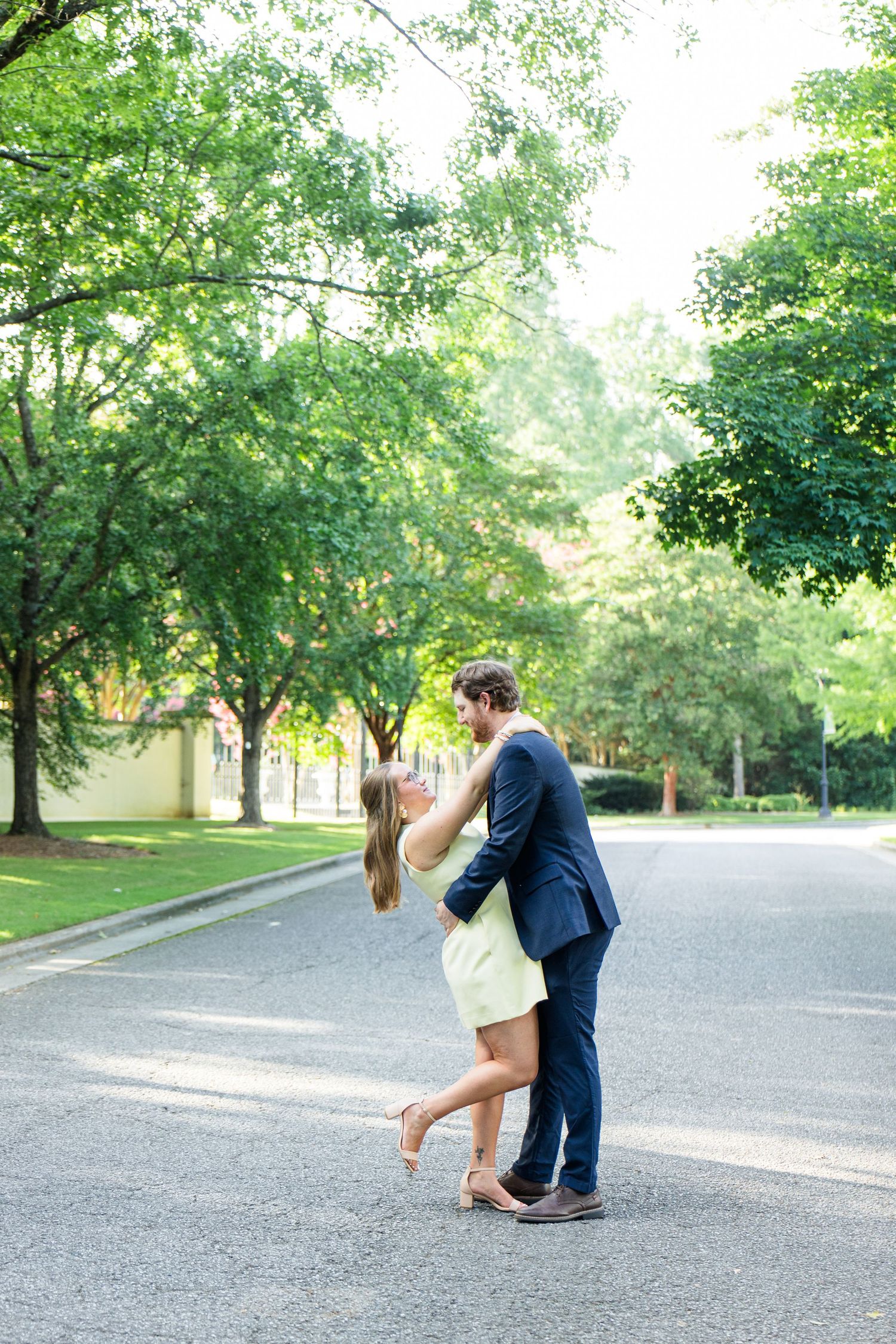 Couple sharing a romantic kiss while dancing on a tree-lined street, woman in yellow dress, man in navy suit.