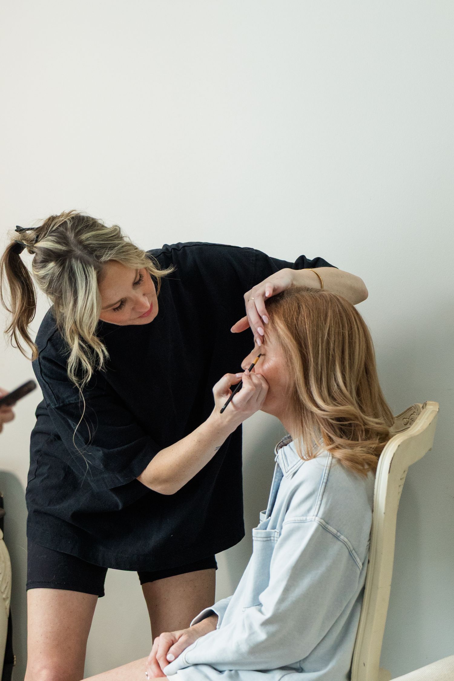 A makeup artist in black applies eye makeup to a seated blonde woman in a light blue top.
