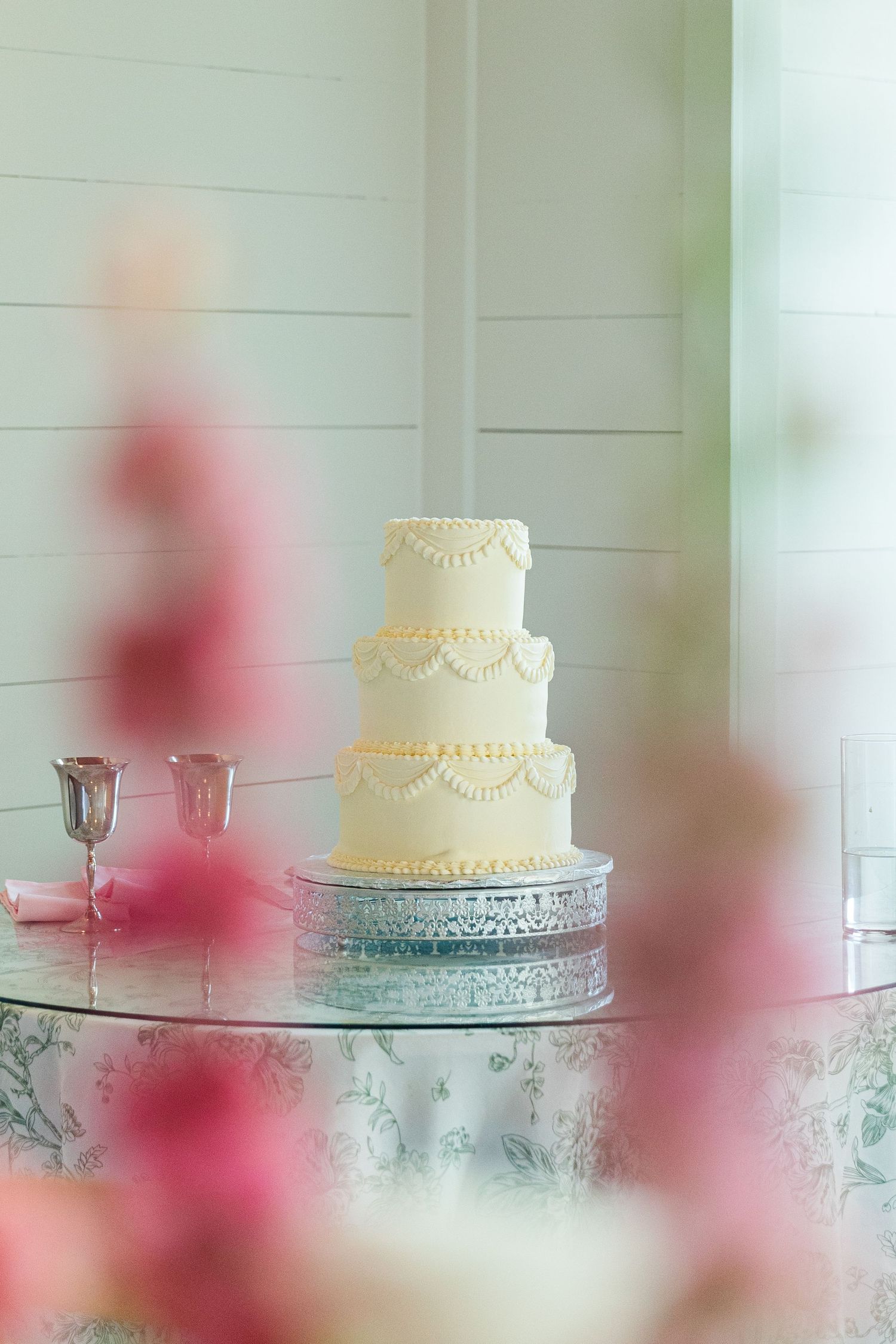 Elegant three-tier white wedding cake with pearl and swag details on a silver stand, surrounded by pink floral decor.