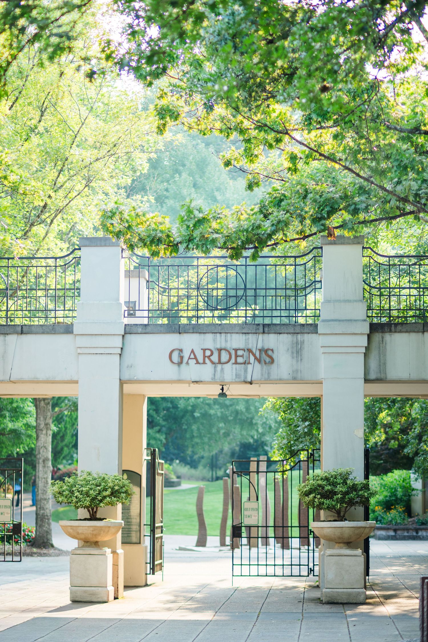 Elegant garden entrance gate with ornamental ironwork and Gardens sign surrounded by trees.