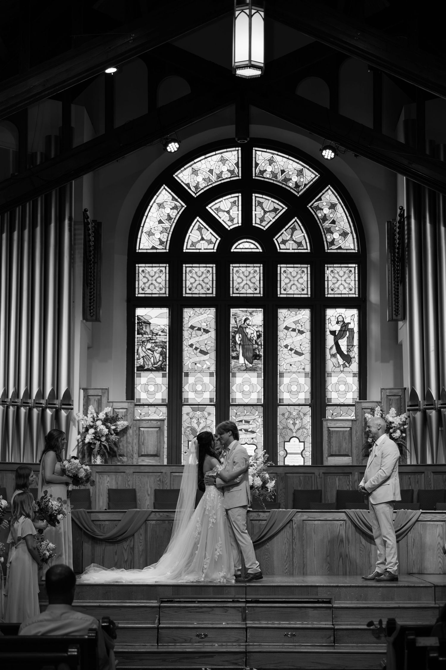 Black and white photo of bride and groom exchanging vows at church altar with stunning stained glass window backdrop.