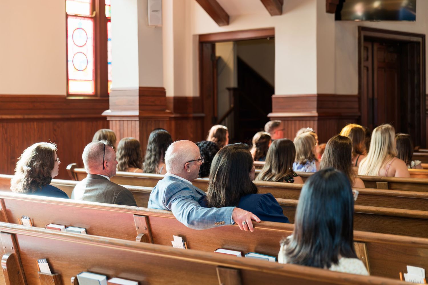 Congregation seated in wooden church pews during a service inside a traditional chapel with stained glass windows.